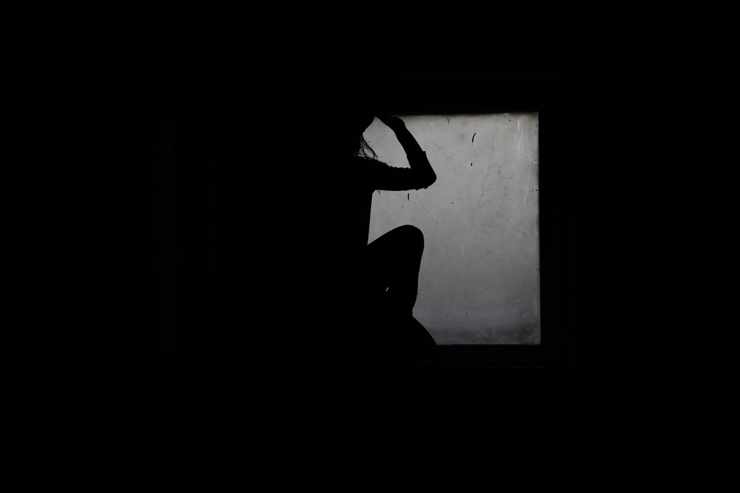Silhouette of a person sitting with one knee up, partially seen in front of a frosted window, with their hand on their head.