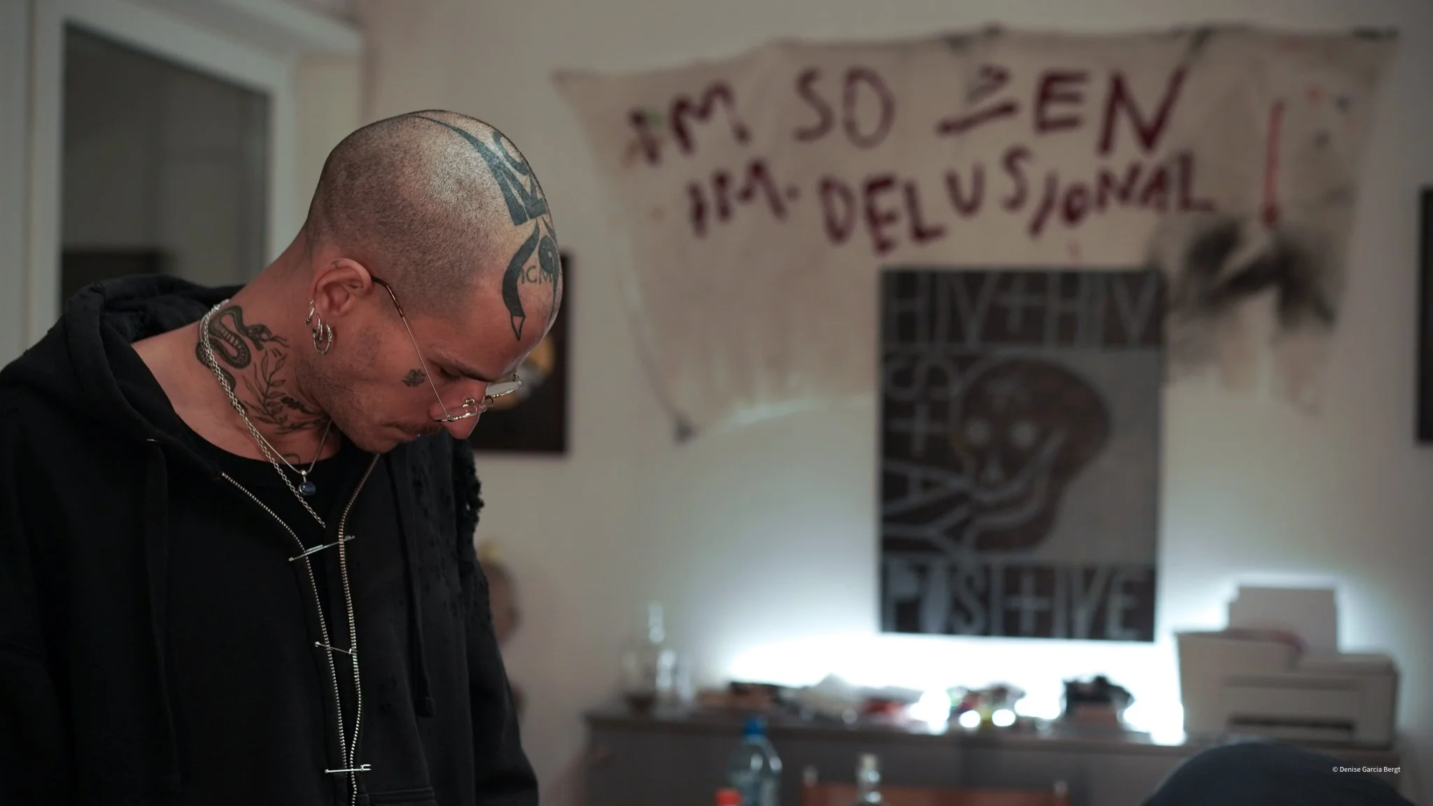 A man with tattoos and piercings, wearing glasses and dressed in black, looks down, in a room with posters and banners on the wall.