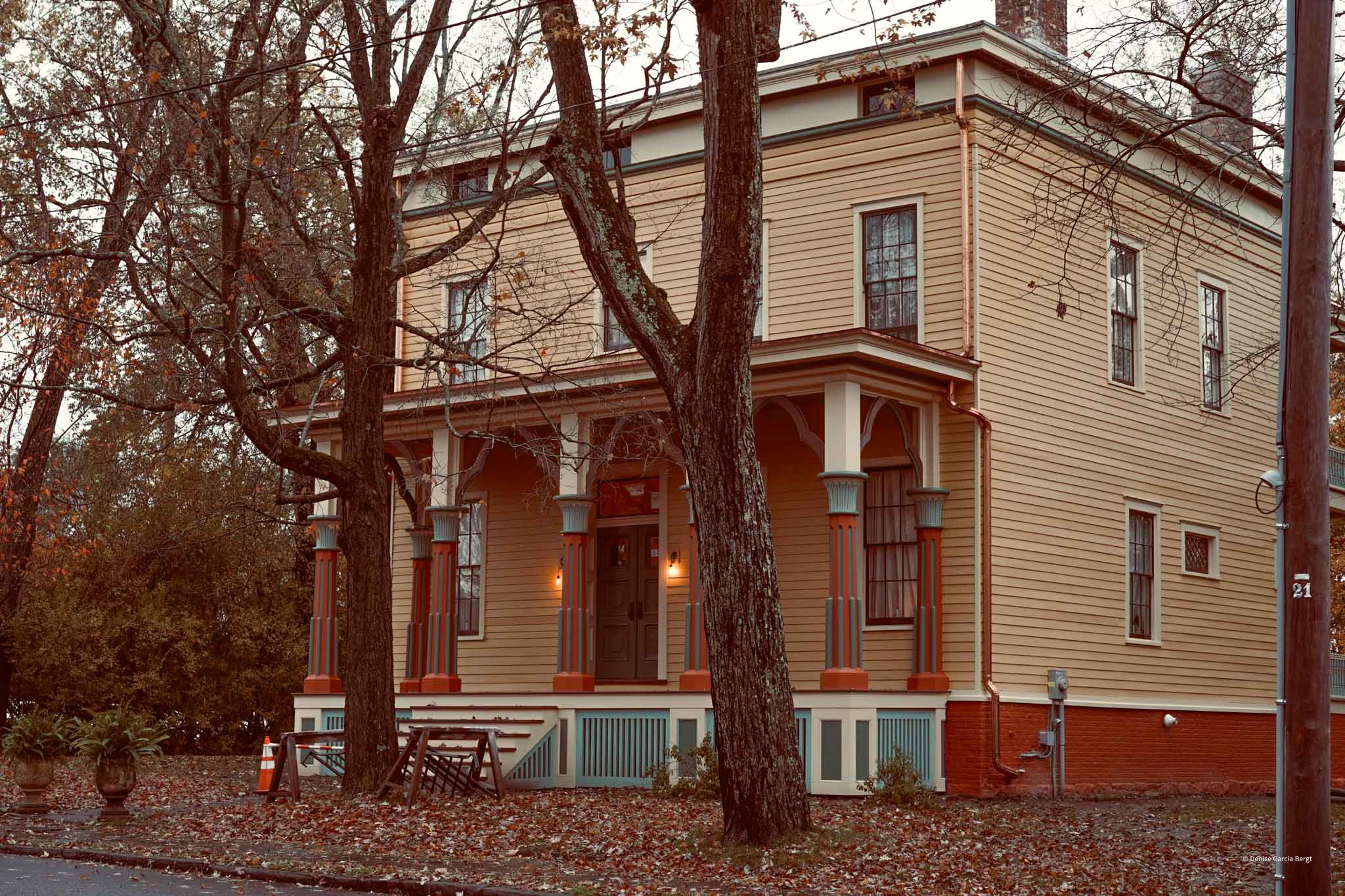 A vintage two-story house with yellow wooden siding, white trim, and a front porch supported by decorative columns. The house is surrounded by leafless trees and fallen autumn leaves on the ground.