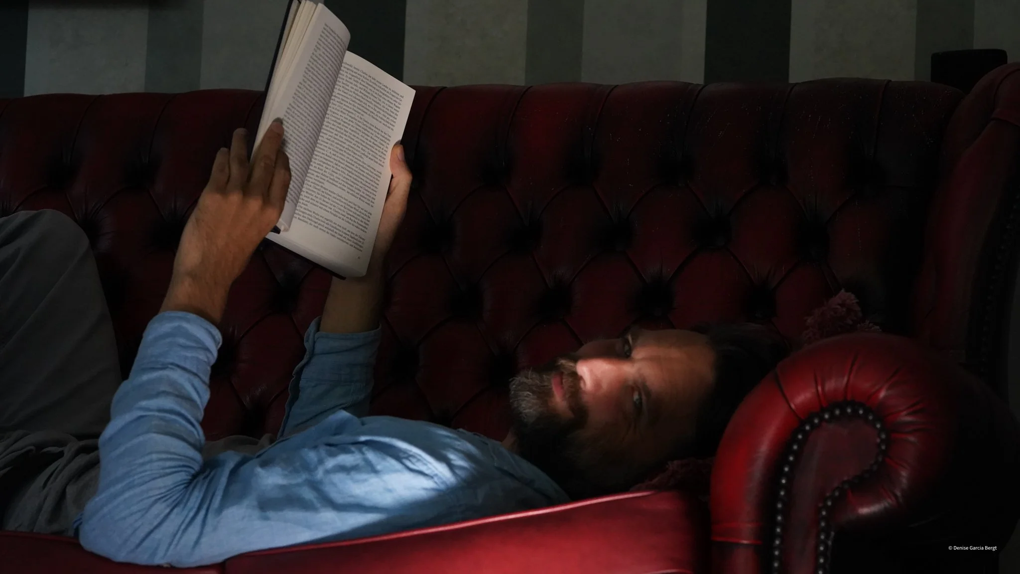 A man with glasses and a beard lies on a red leather tufted sofa, reading a book.