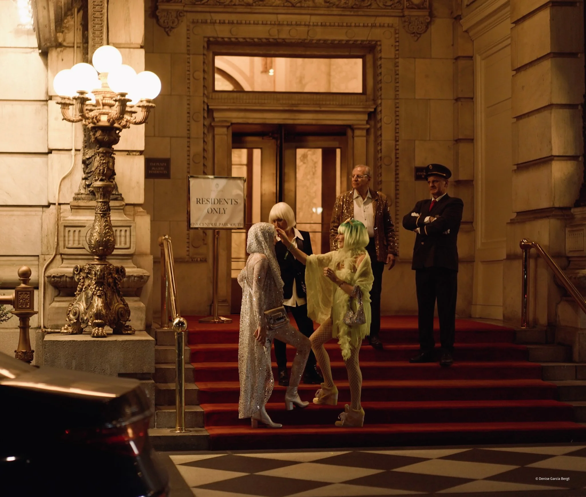 Group of five people standing on a red carpet staircase outside a historic building at night, dressed in fashionable clothing, with one person in a shiny silver outfit with a hood, and others in colorful costumes, engaged in conversation.