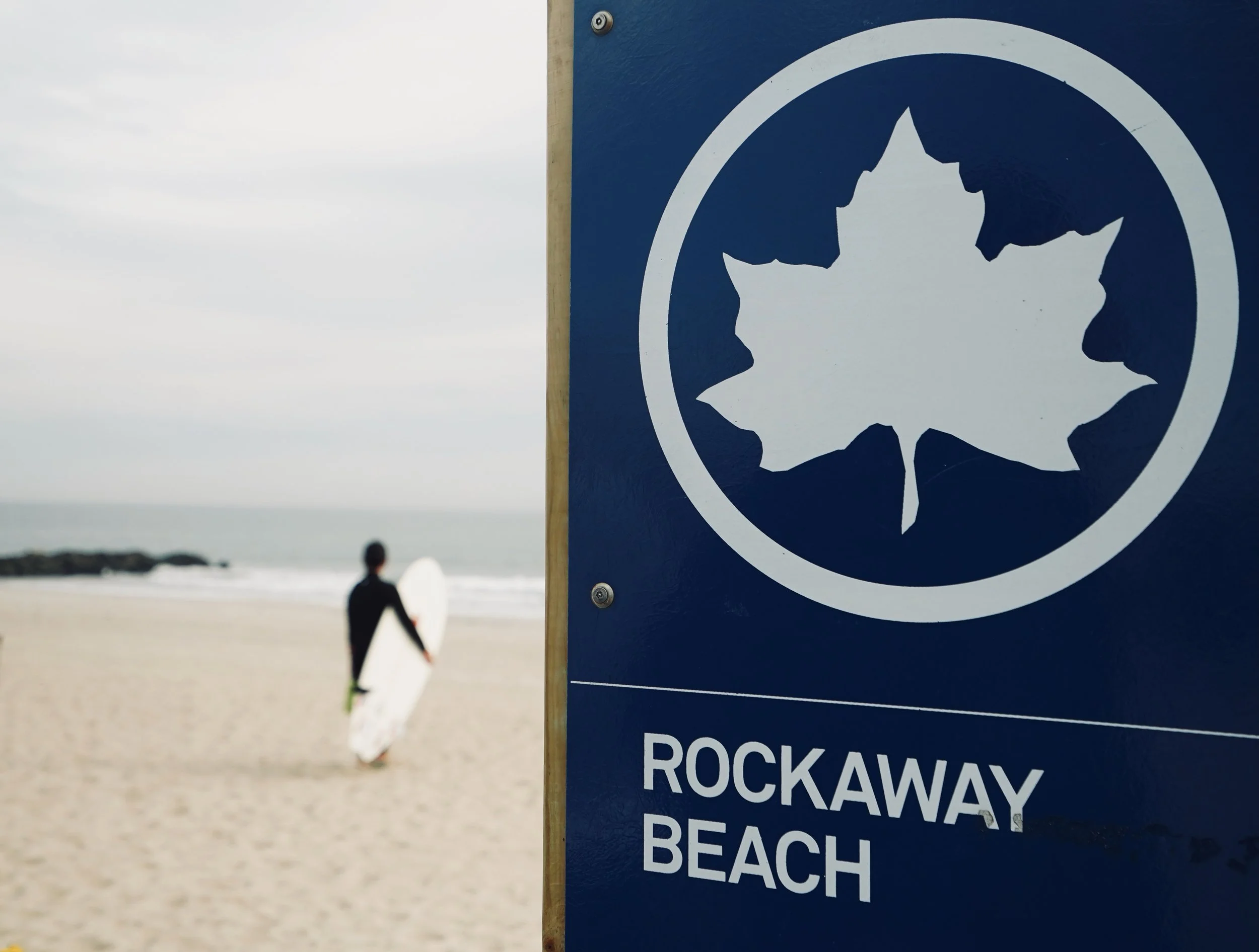 Sign for Rockaway Beach with a maple leaf symbol, with a person holding a surfboard walking on the sandy beach in the background under an overcast sky.