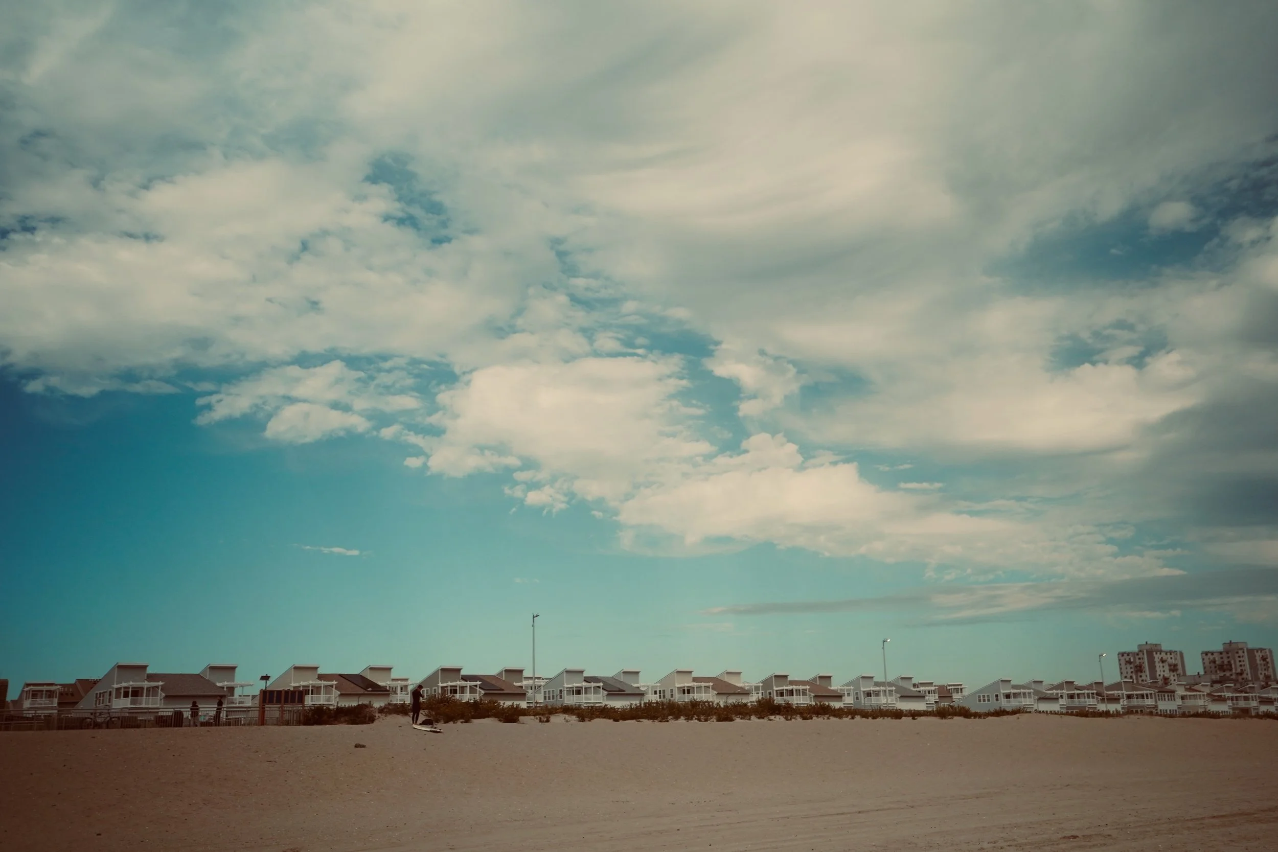 Beach with sand and row of houses in the distance under partly cloudy sky.