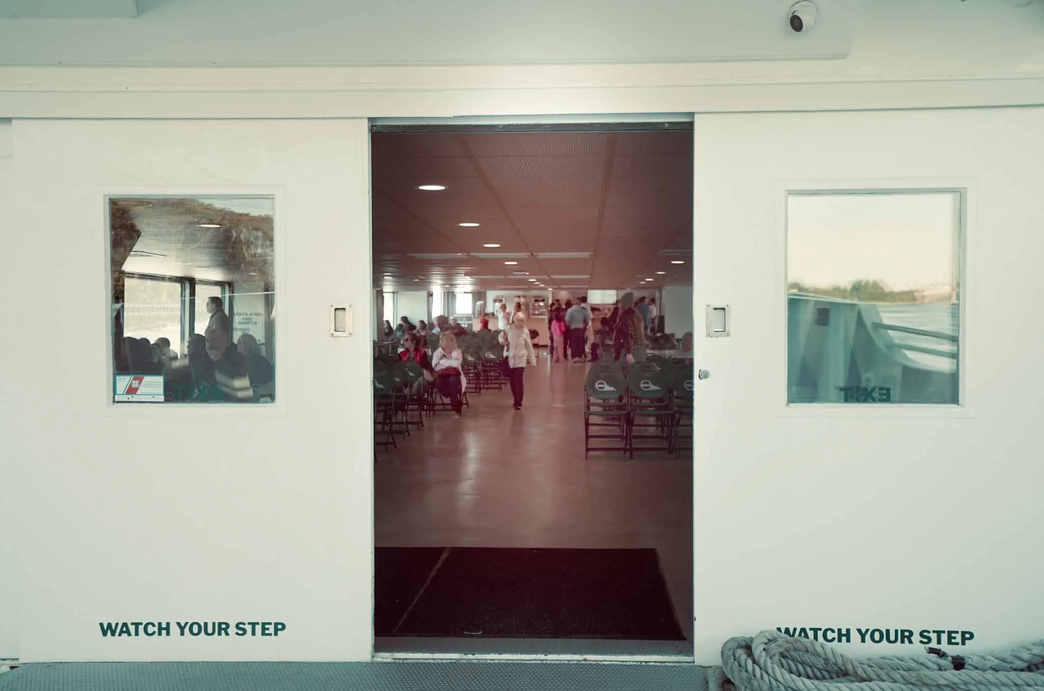 View of an airport terminal through open sliding doors, with people waiting and walking inside, and chairs arranged in rows.