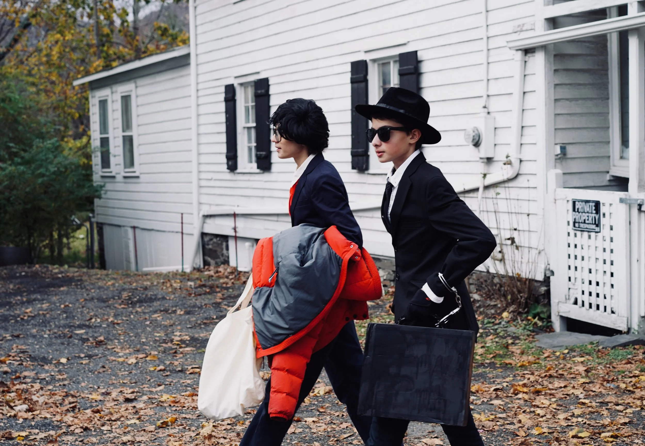 Two women dressed in black suits with sunglasses and accessories walking past a white house with black shutters, carrying bags and a trunk, autumn leaves on the ground.
