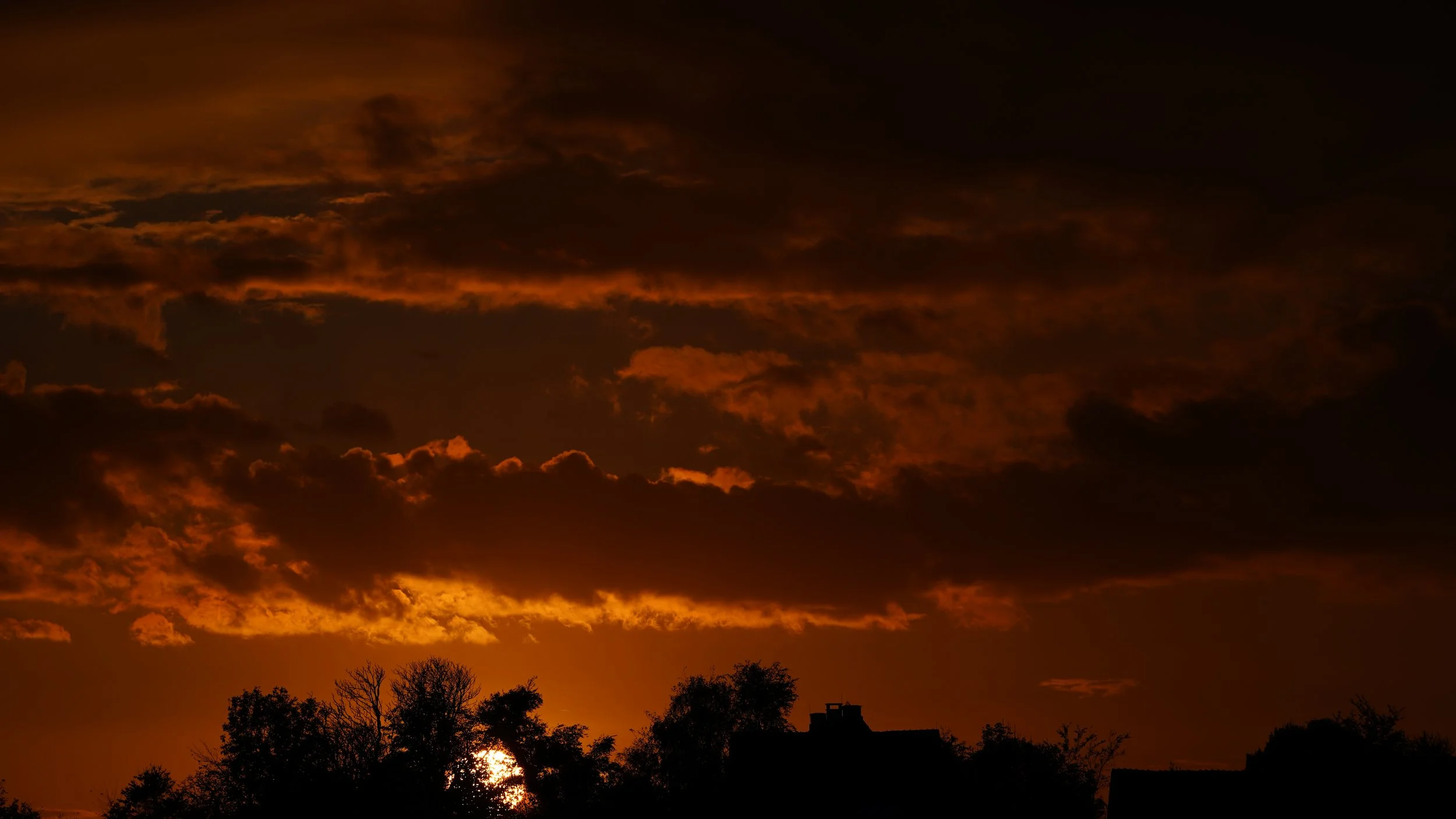 Sunset with orange and dark clouds over silhouette trees and buildings.