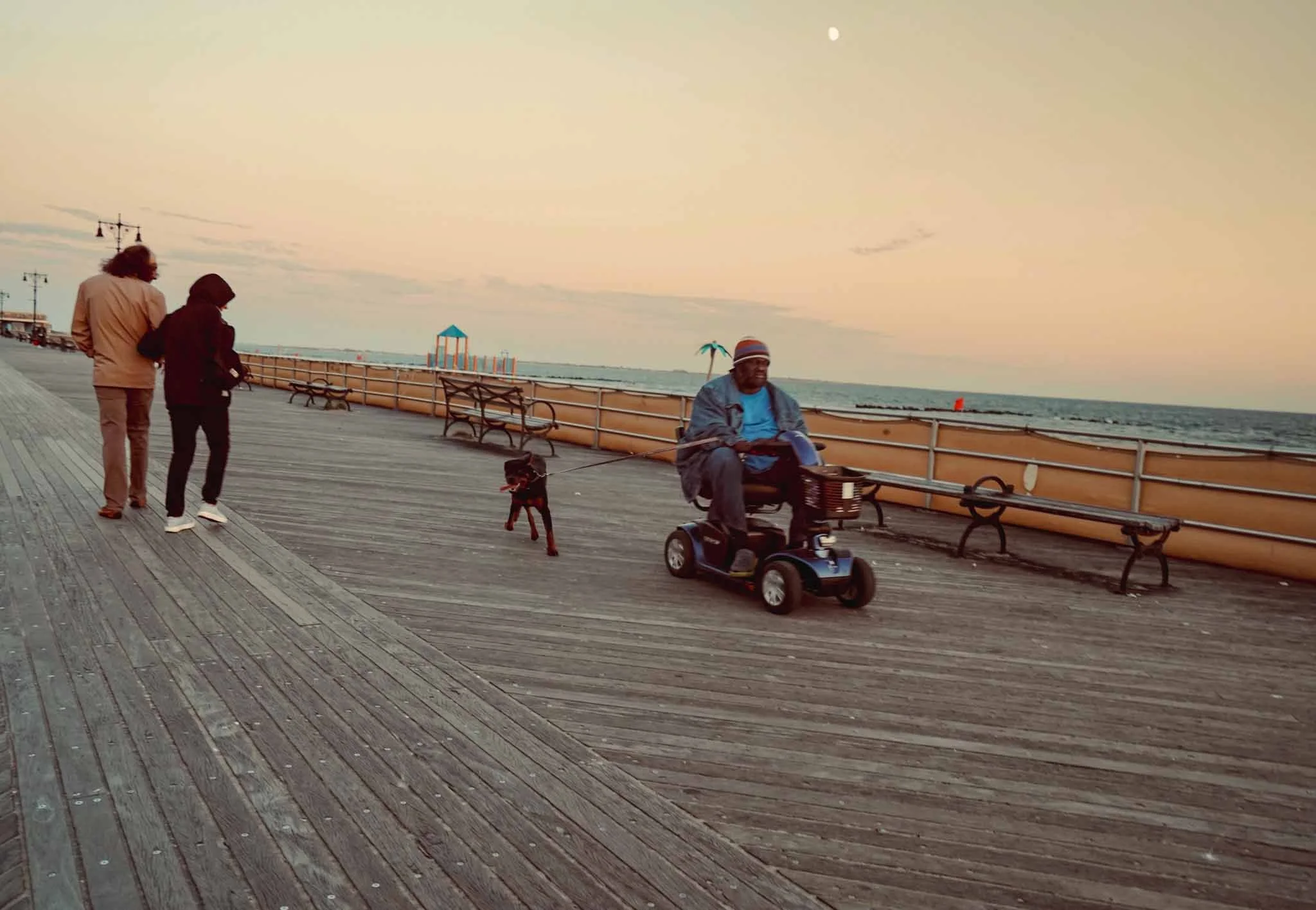 People walking along a wooden pier by the ocean at sunset, with benches, a large man on a mobility scooter, and a dog.