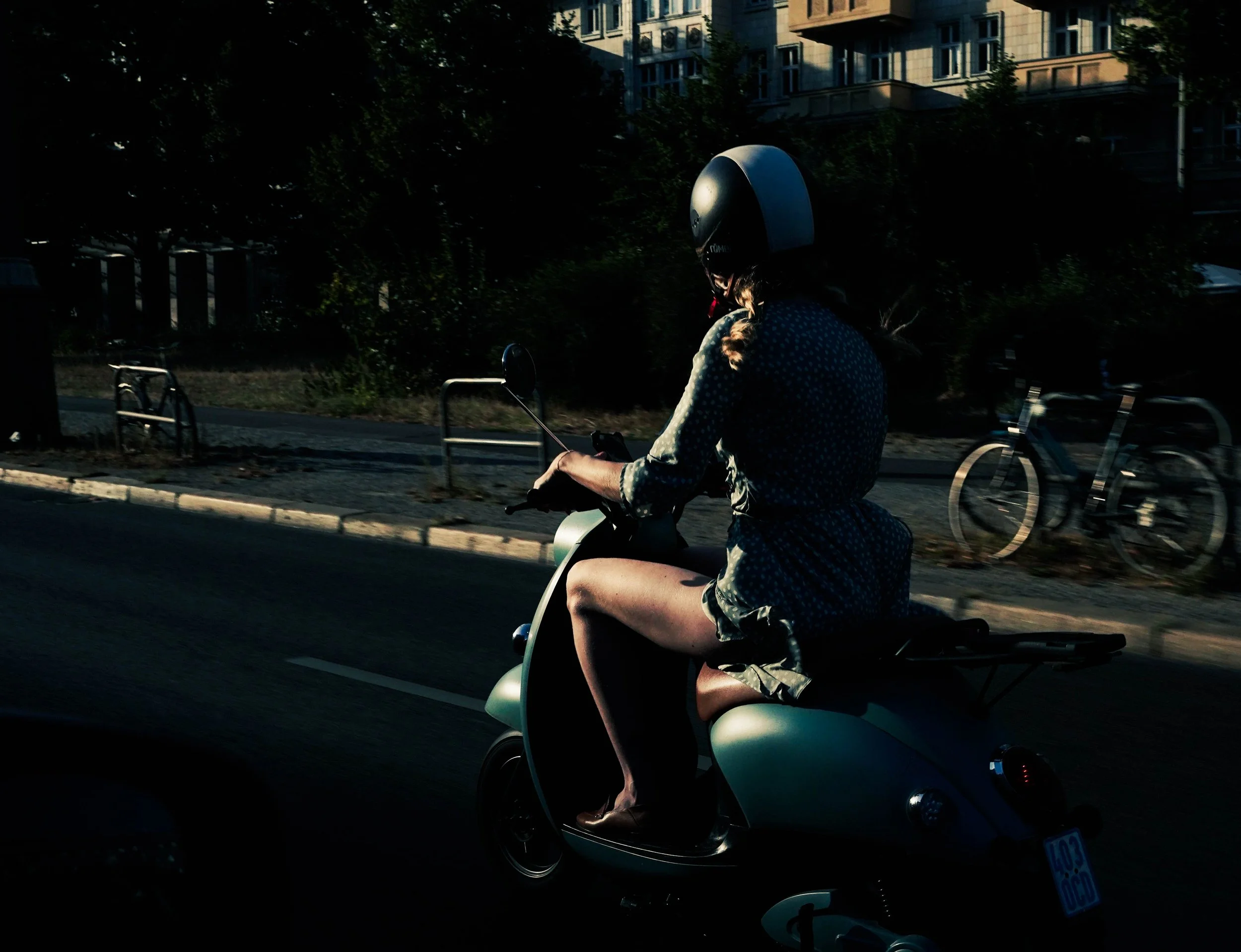 Person riding a scooter wearing a helmet and a dress, with bicycles parked on the sidewalk and trees in the background.
