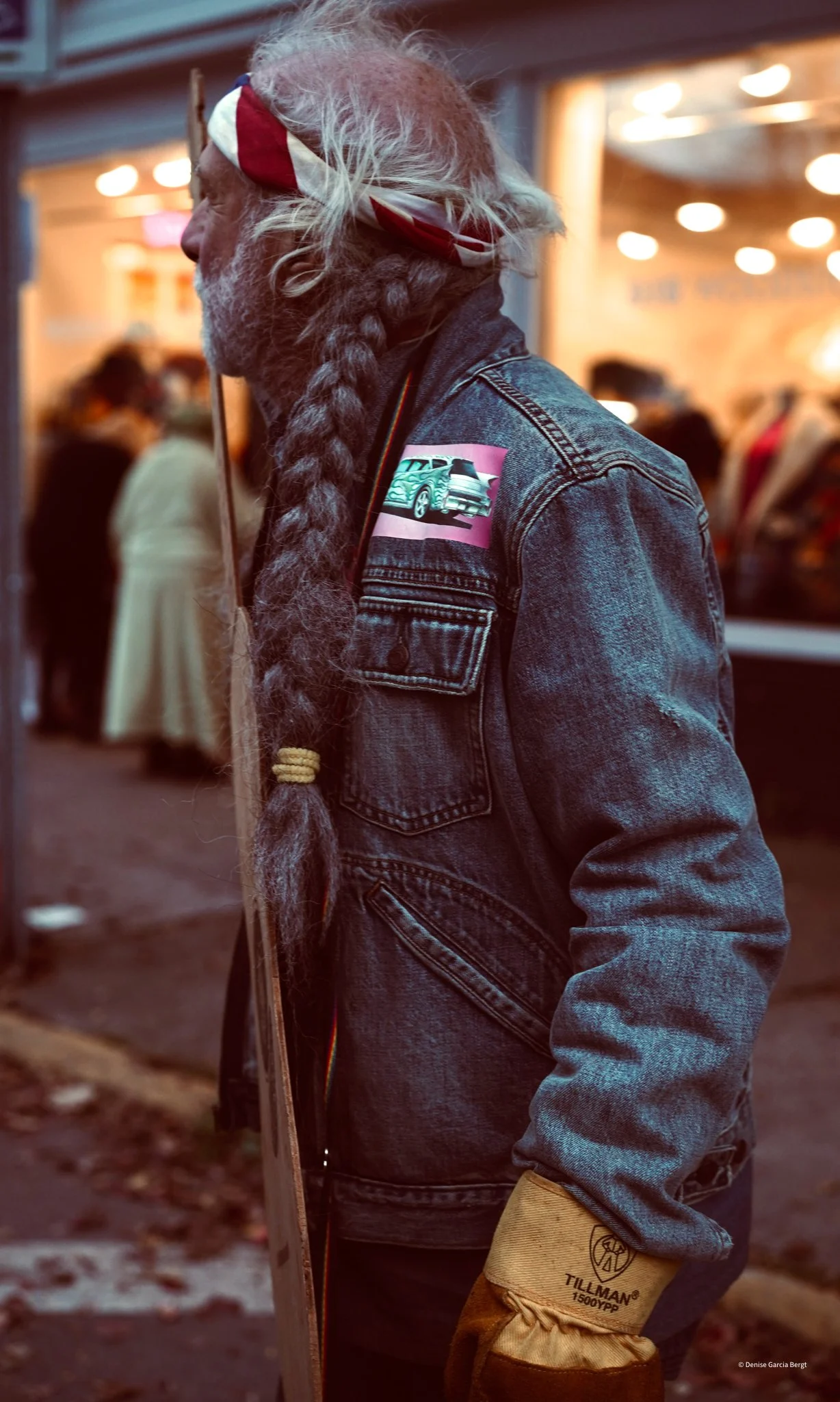 Side view of a bearded man with long hair in a braid, wearing a denim jacket, a red and white bandana, and tan work gloves, standing outside at night.