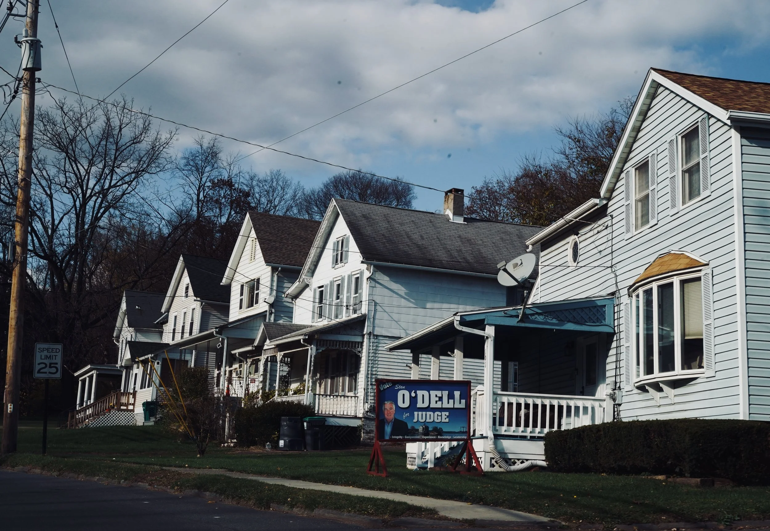 A row of white residential houses with porches, satellite dishes, and trees in the background, with a political campaign sign for O'Dell Judge in the foreground.