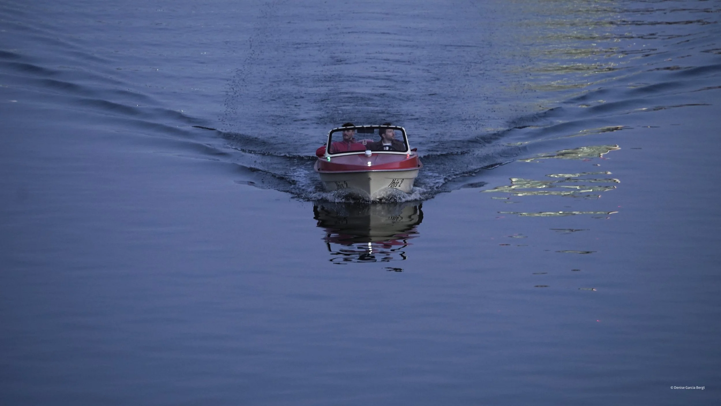 A small red and white motorboat with two men onboard, moving across a calm body of water, creating ripples behind it.