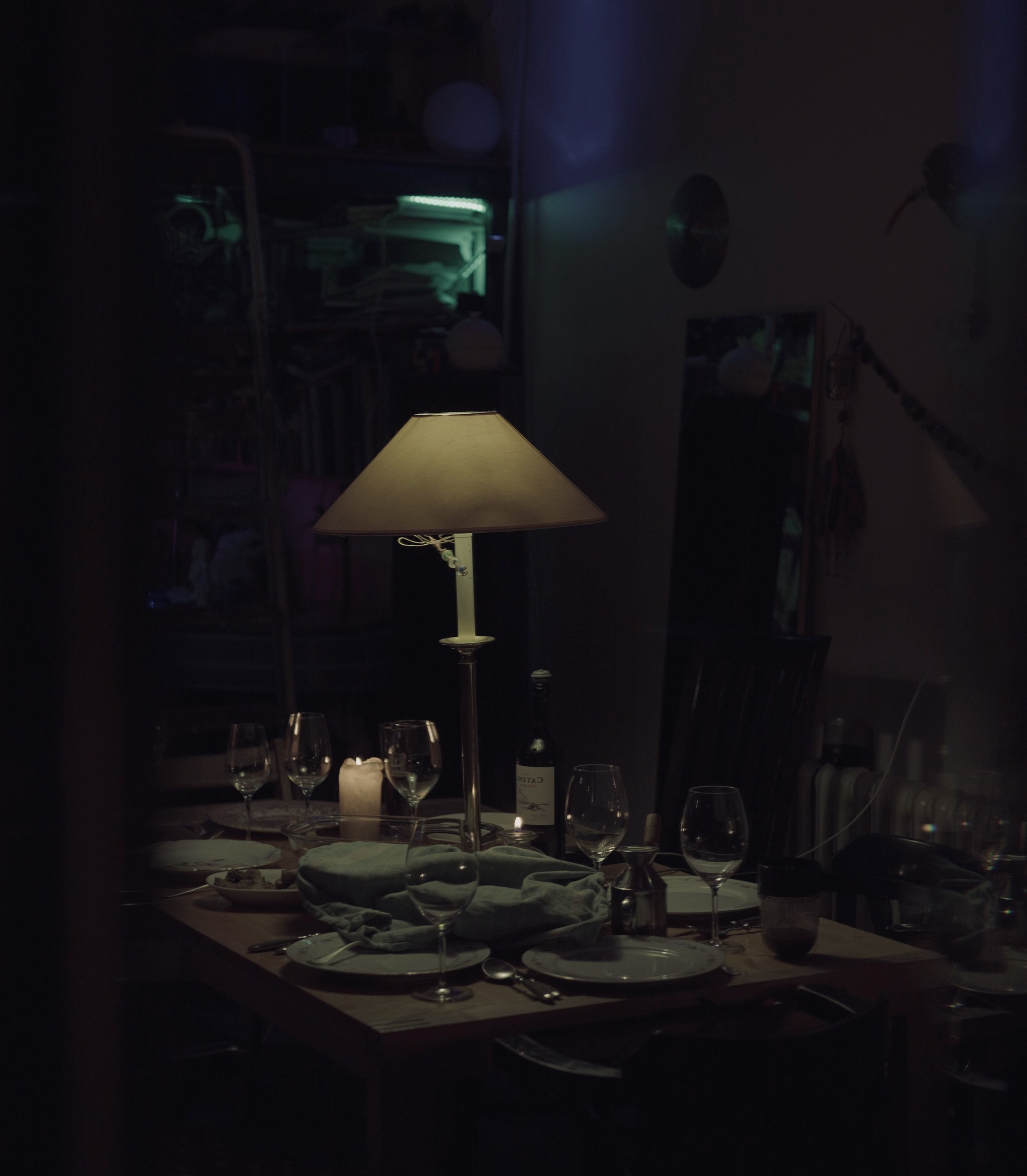 Dining table set with wine glasses, a bottle of wine, a candle, and a table lamp in a dimly lit room.