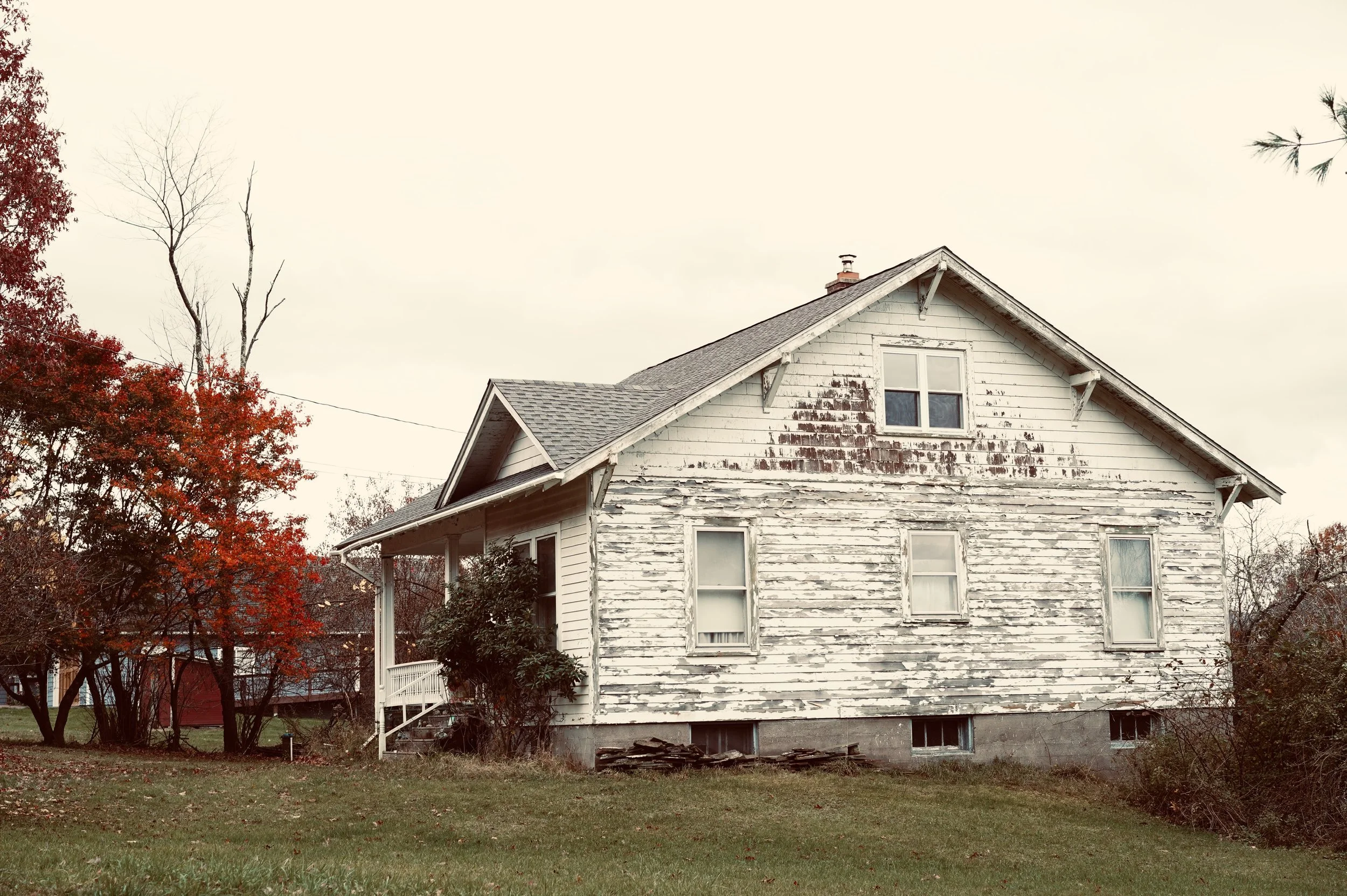 An old, weathered white house with peeling paint, surrounded by trees with red leaves, situated on a grassy lot.