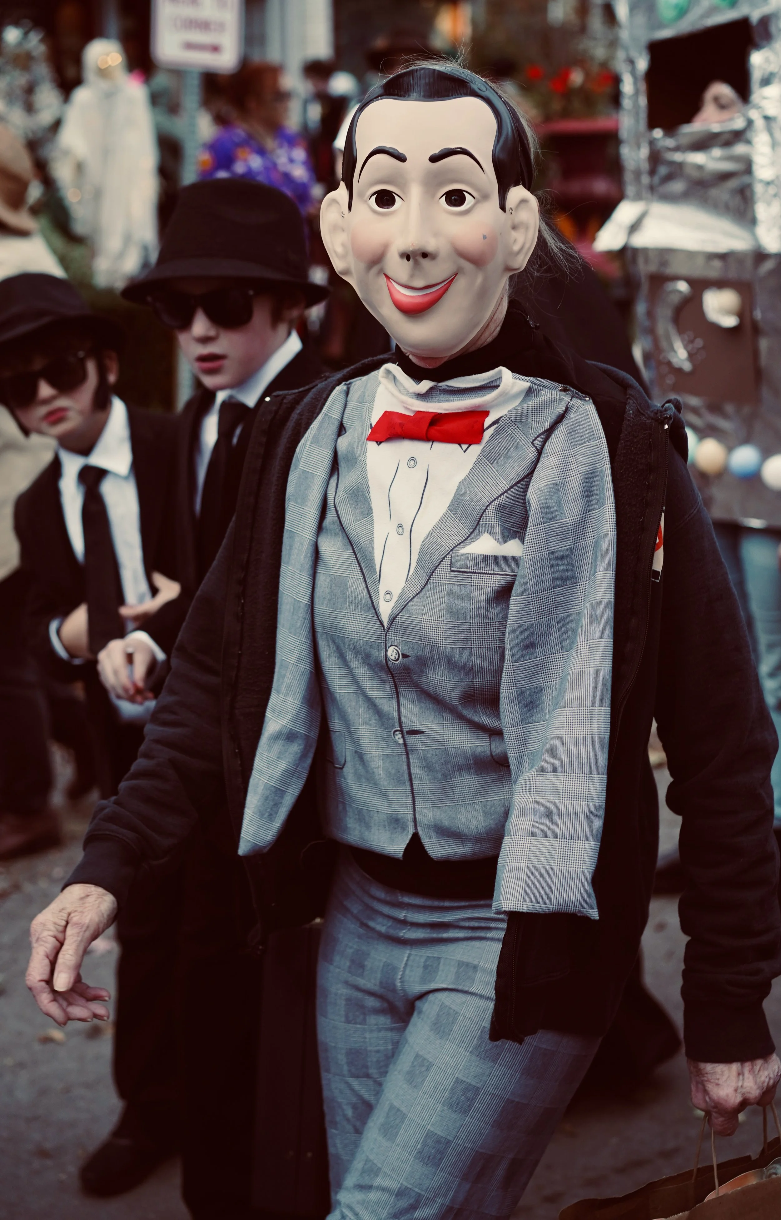 Person dressed in a cartoonish Adlai Stevenson costume with a mask of a smiling man, wearing a plaid suit, red bow tie, and gray plaid pants, at an outdoor event in daytime.