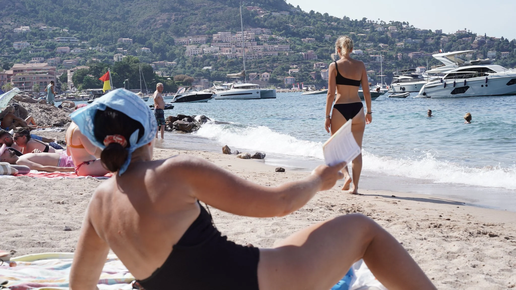 Beach scene with people sunbathing, swimming, and walking near boats and yachts at a marina, with hills and houses in the background.