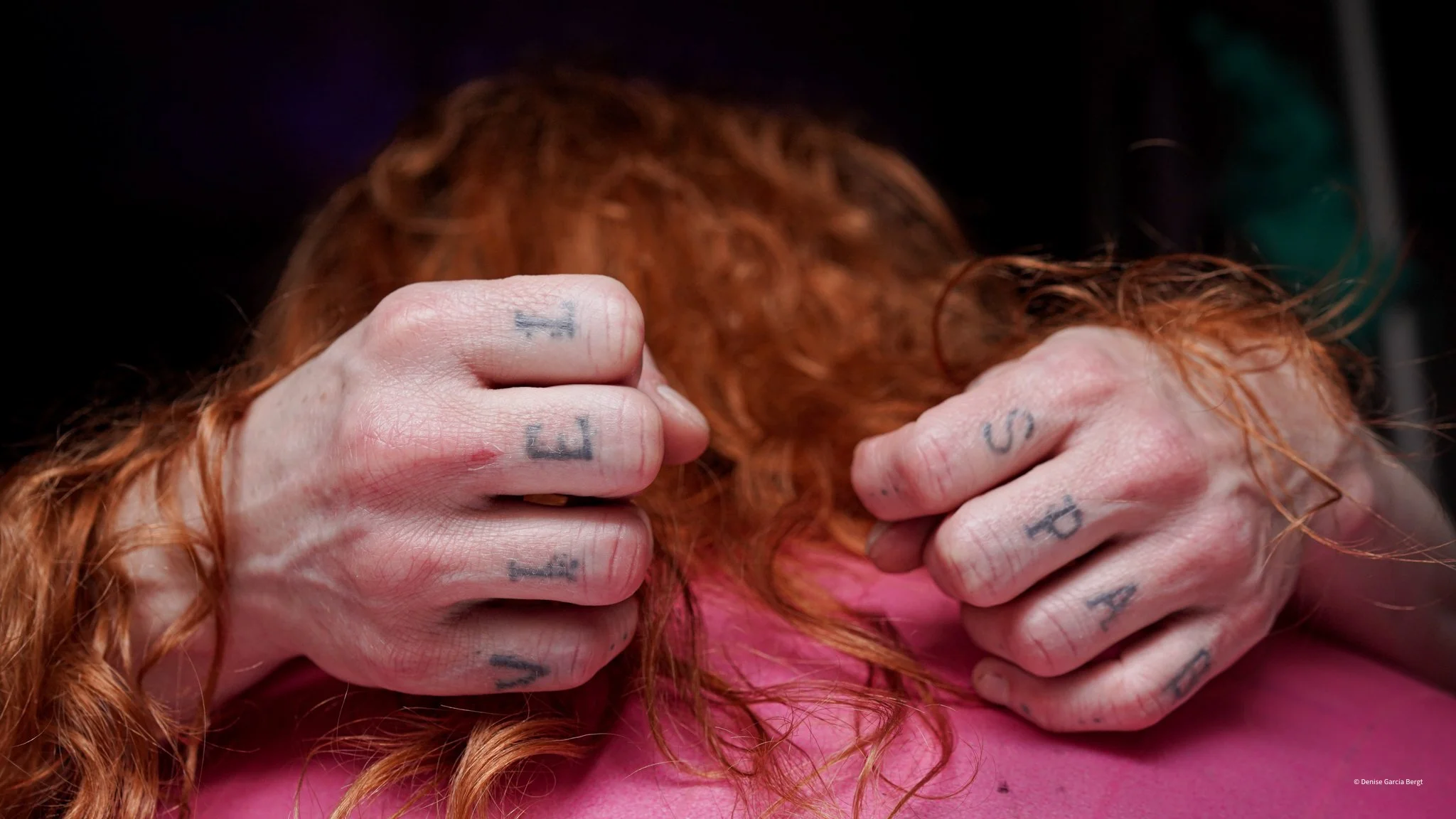 Close-up of a person's hands with tattoos gripping their red hair, which is resting on a pink surface.