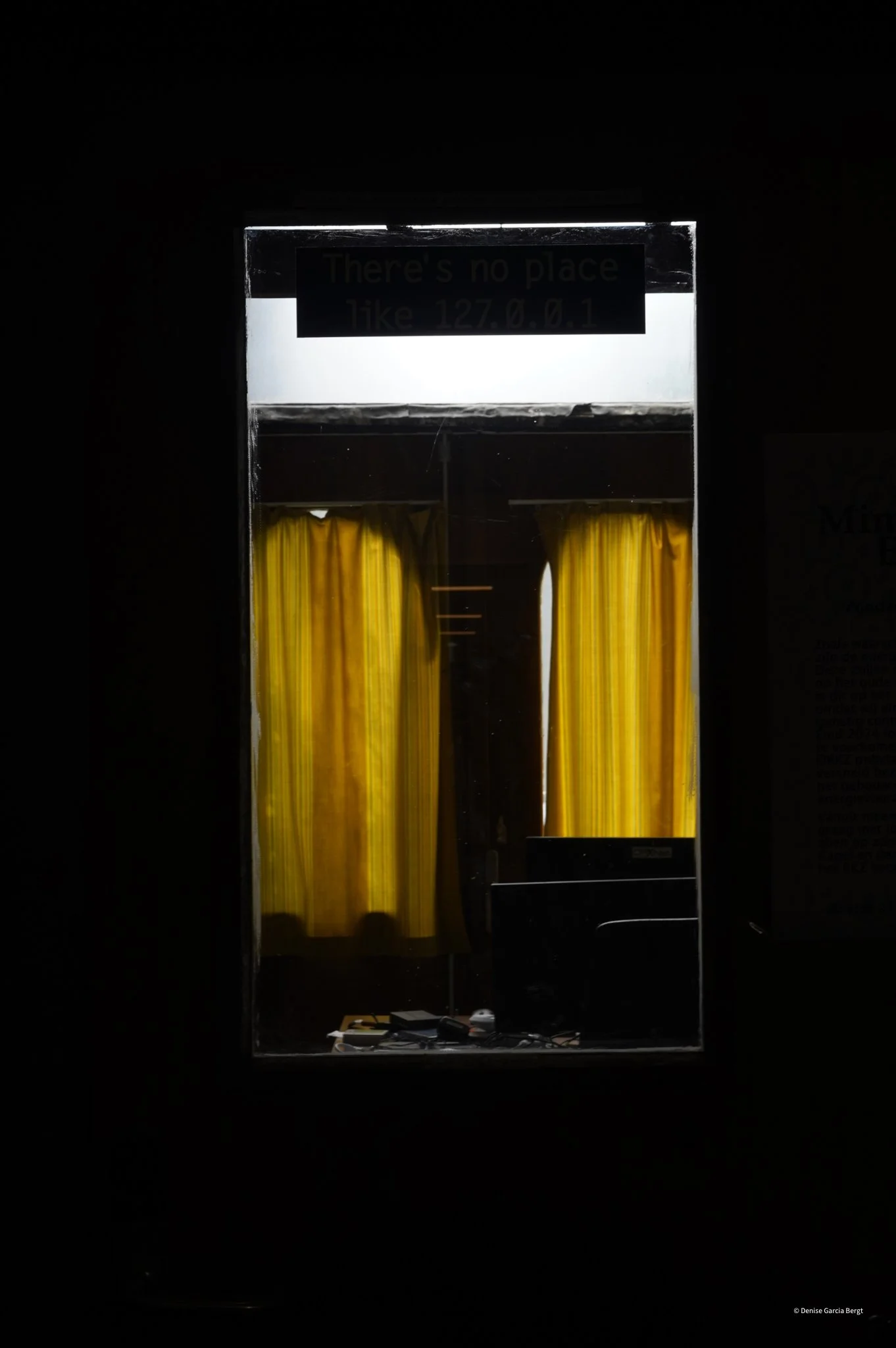 A reflection of a window with yellow curtains and a sign that reads 'There's no place like 127.0.0.1' in a dark room, with a cluttered desk or surface at the bottom containing various objects.