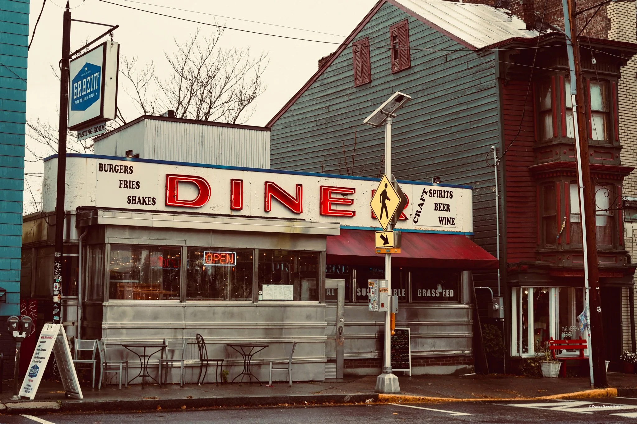 A vintage roadside diner with a neon sign spelling 'DINE'; signs indicate it serves burgers, fries, shakes, craft spirits, beer, and wine. The diner has a metal exterior, large windows with neon 'OPEN' sign, outdoor seating, and a red awning, with a 