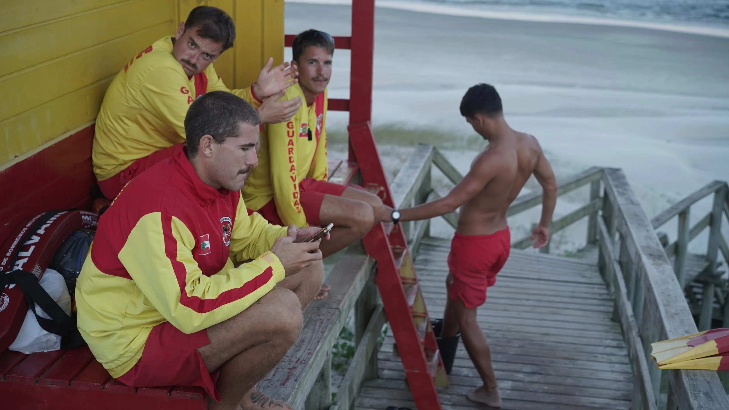 Four lifeguards sitting on a wooden structure near the beach, one of them using a smartphone, while a shirtless young man in red shorts stands on the stairs leading into the ocean, which has waves in the background.