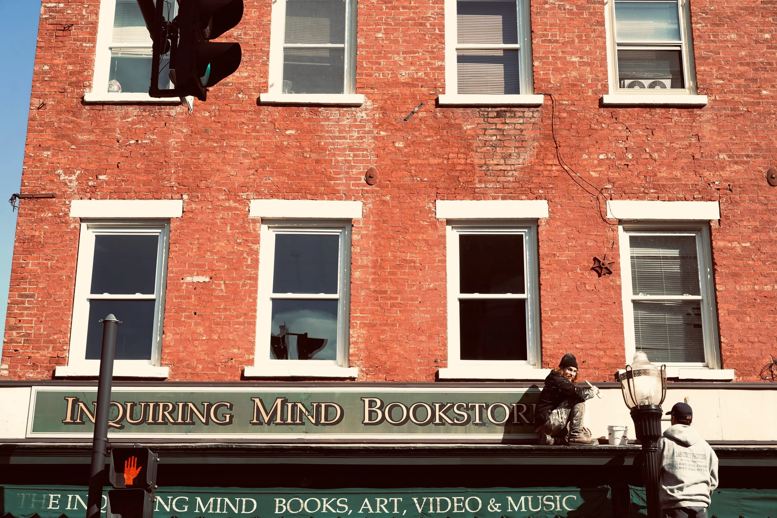 A brick building with six windows and a sign for Quiring Mind Bookstore. Two people are on the roof, one sitting and the other standing, near a large street lamp. A traffic light shows a red hand signal.