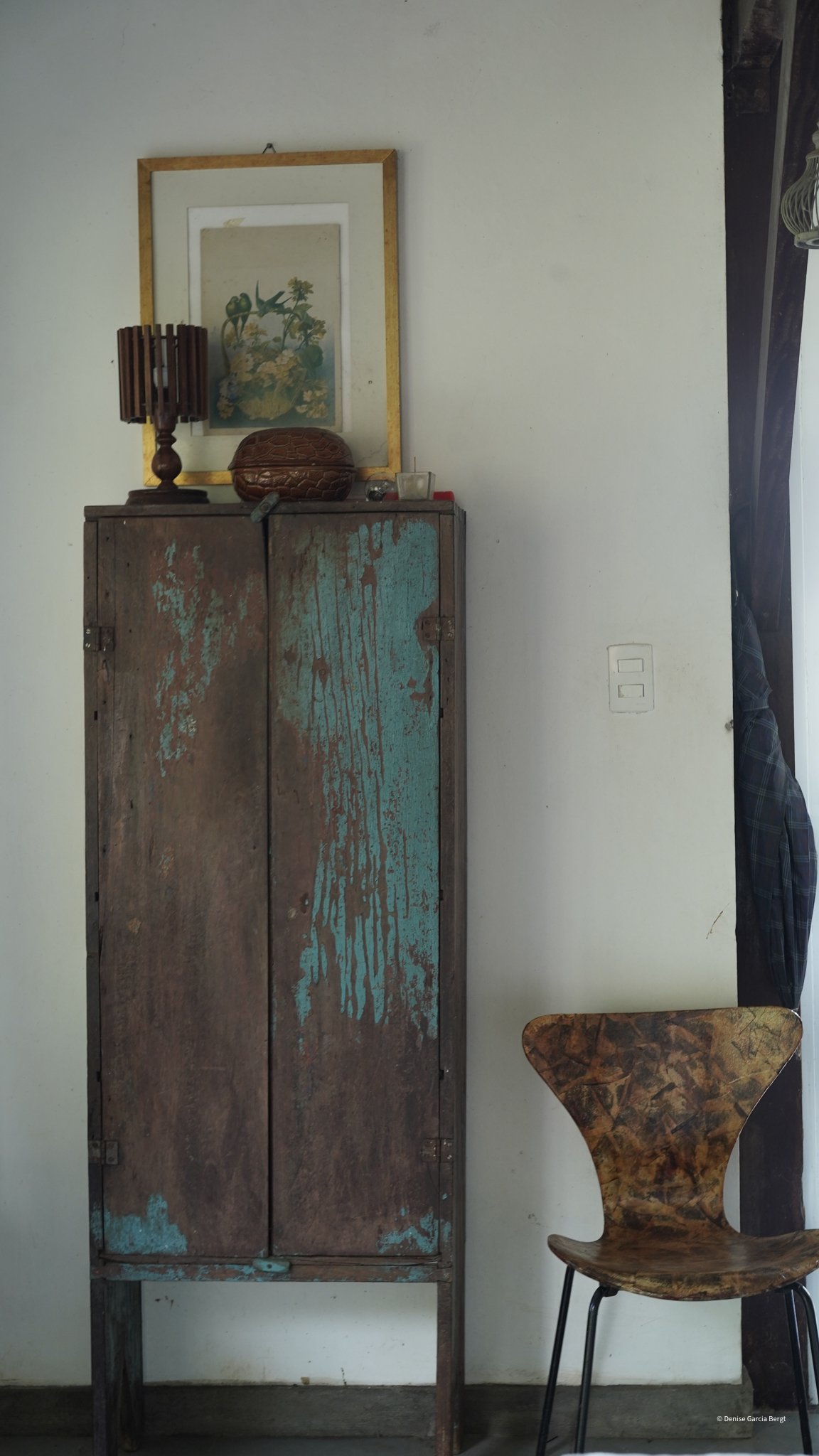 A rustic wooden cabinet with distressed paint, a framed botanical print above, a table lamp, and small decorative objects on top, with a modern wooden chair beside it.