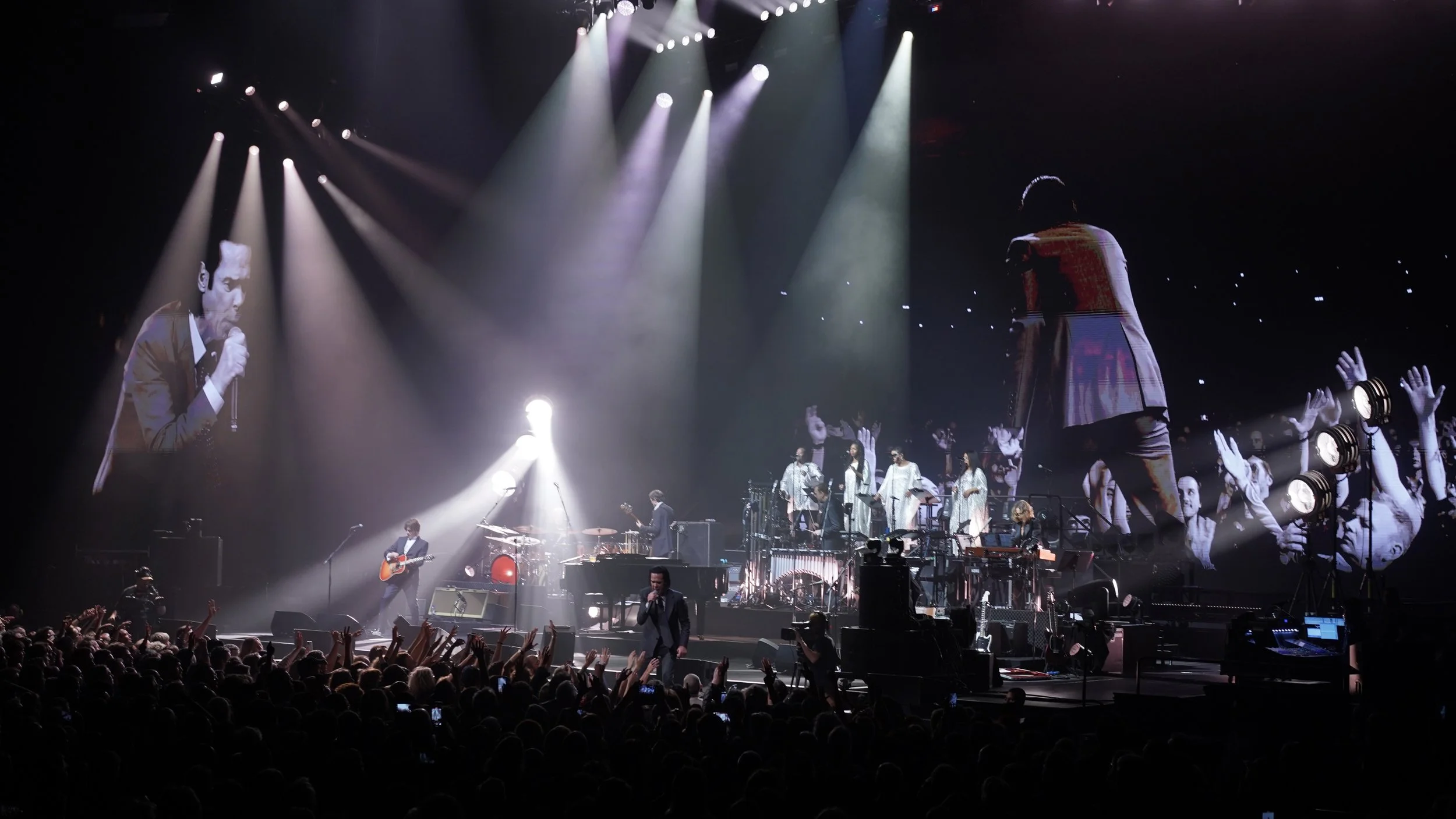 A live concert with a band performing on stage, featuring multiple musicians and singers, illuminated by stage lights. There is a large screen projecting images of some performers, and the audience is visible with many people raising their hands.