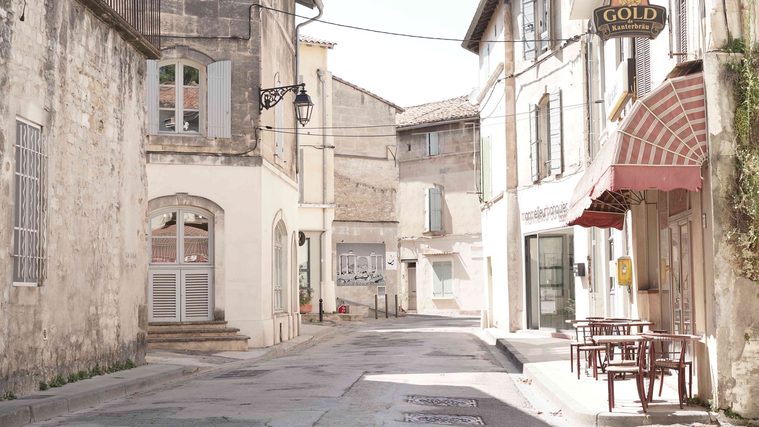 A quiet European street with old stone buildings, some with shutters, and outdoor seating for a café or restaurant with red chairs and tables under a canopy.