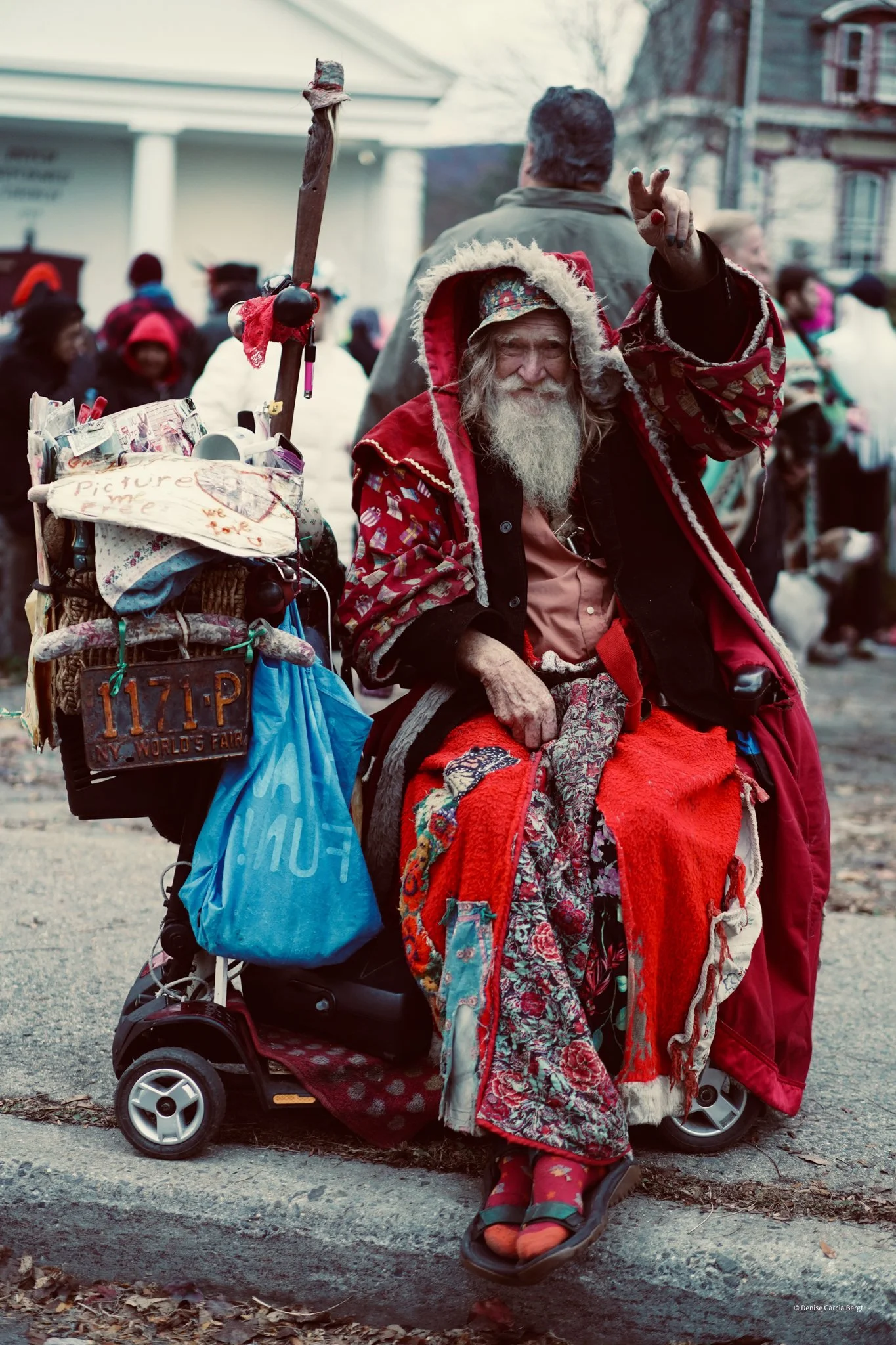 An elderly man dressed as Santa Claus sitting in a mobility scooter, waving. He is wearing a red hoodie with a beige lining, and layered red and patterned clothes. His scooter is heavily decorated with bags, signs, and personal belongings, indicating