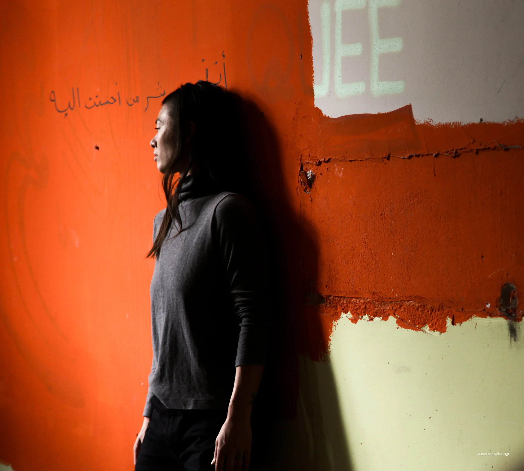 A woman with dark hair leaning against a textured orange wall with handwriting and a projection of text.