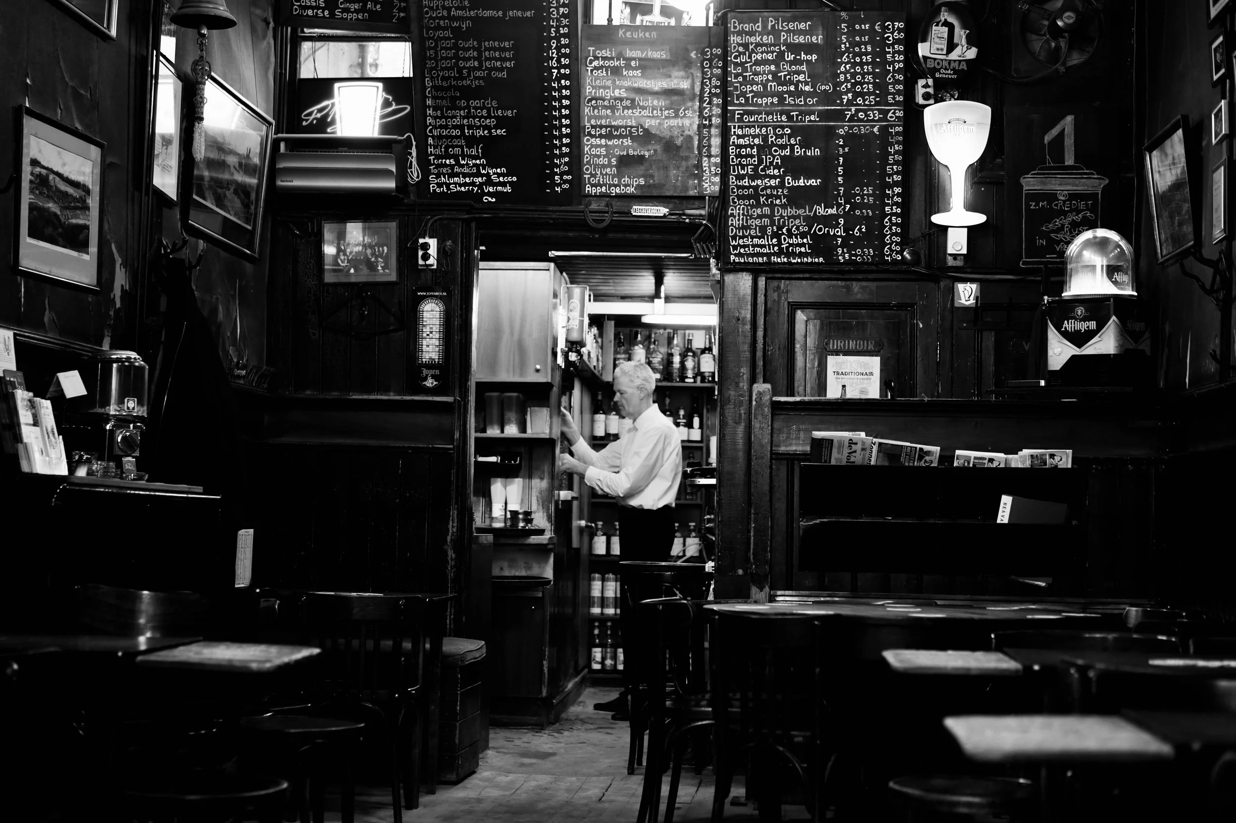 Black and white photo of a bar or cafe interior with empty tables and chairs. A man with white hair and a white shirt stands behind the bar, organizing shelves. The background features a chalkboard menu with various drink options, framed pictures on 