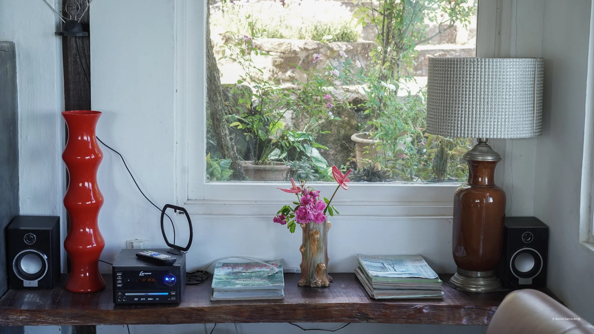 A window with a view of outdoor plants and a stone wall, a wooden table with a red vase, a brown lamp, a small black radio, a stack of magazines, and a floral arrangement in a wooden vase.