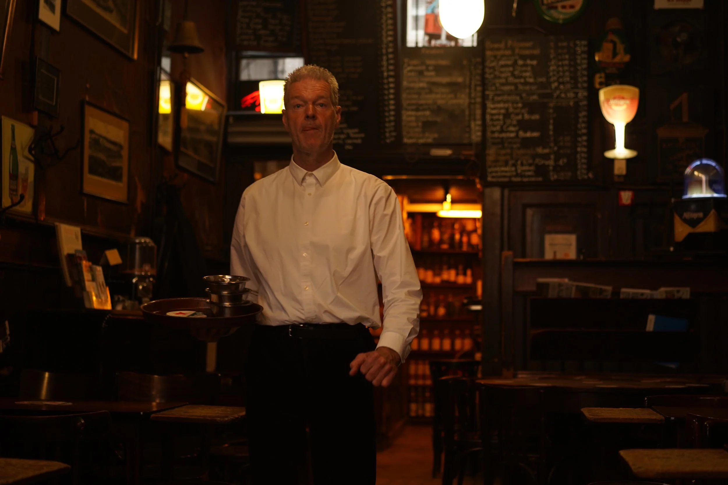 A man in a white shirt standing inside a dimly lit restaurant, holding a tray with items on it, with a menu board and various decorations on the walls in the background.