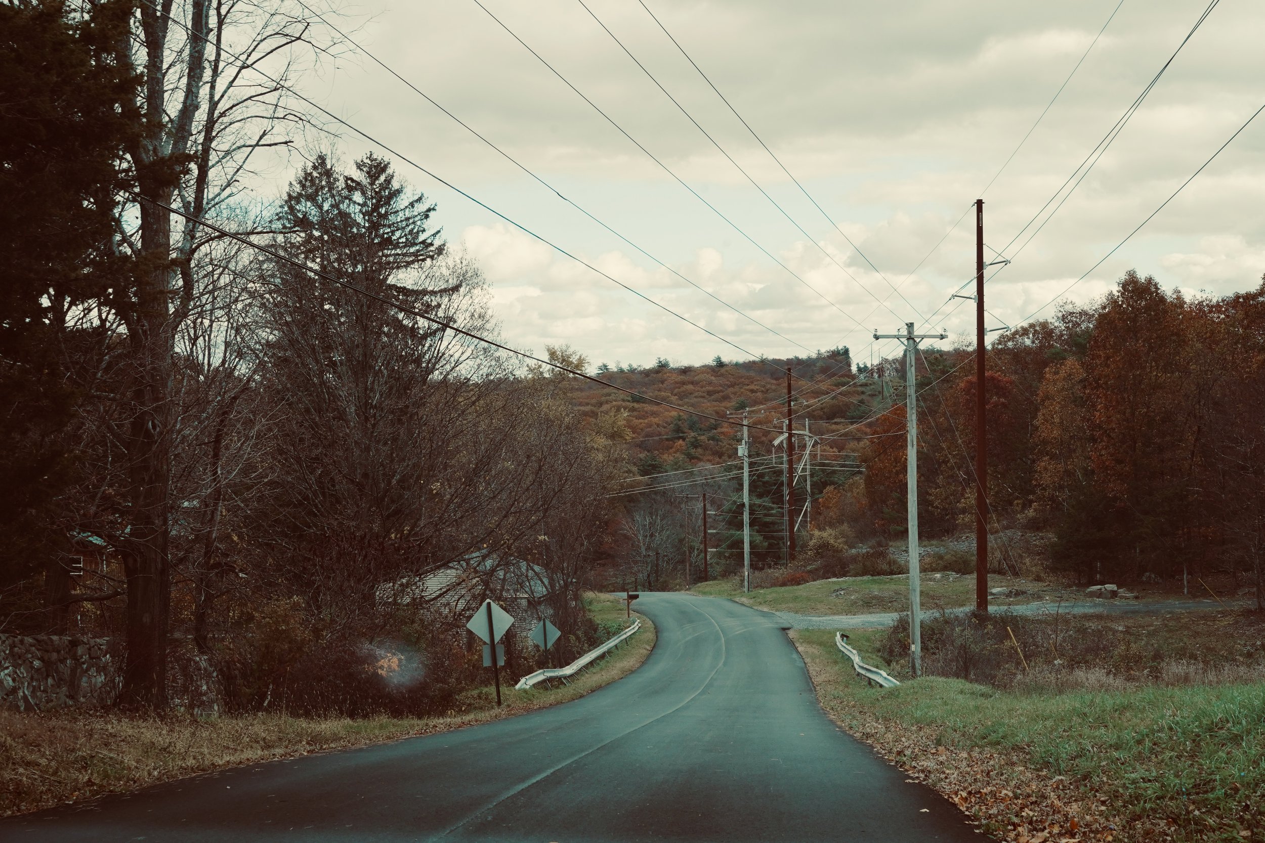 A winding rural road with utility poles and power lines, surrounded by trees with autumn foliage, under a cloudy sky.