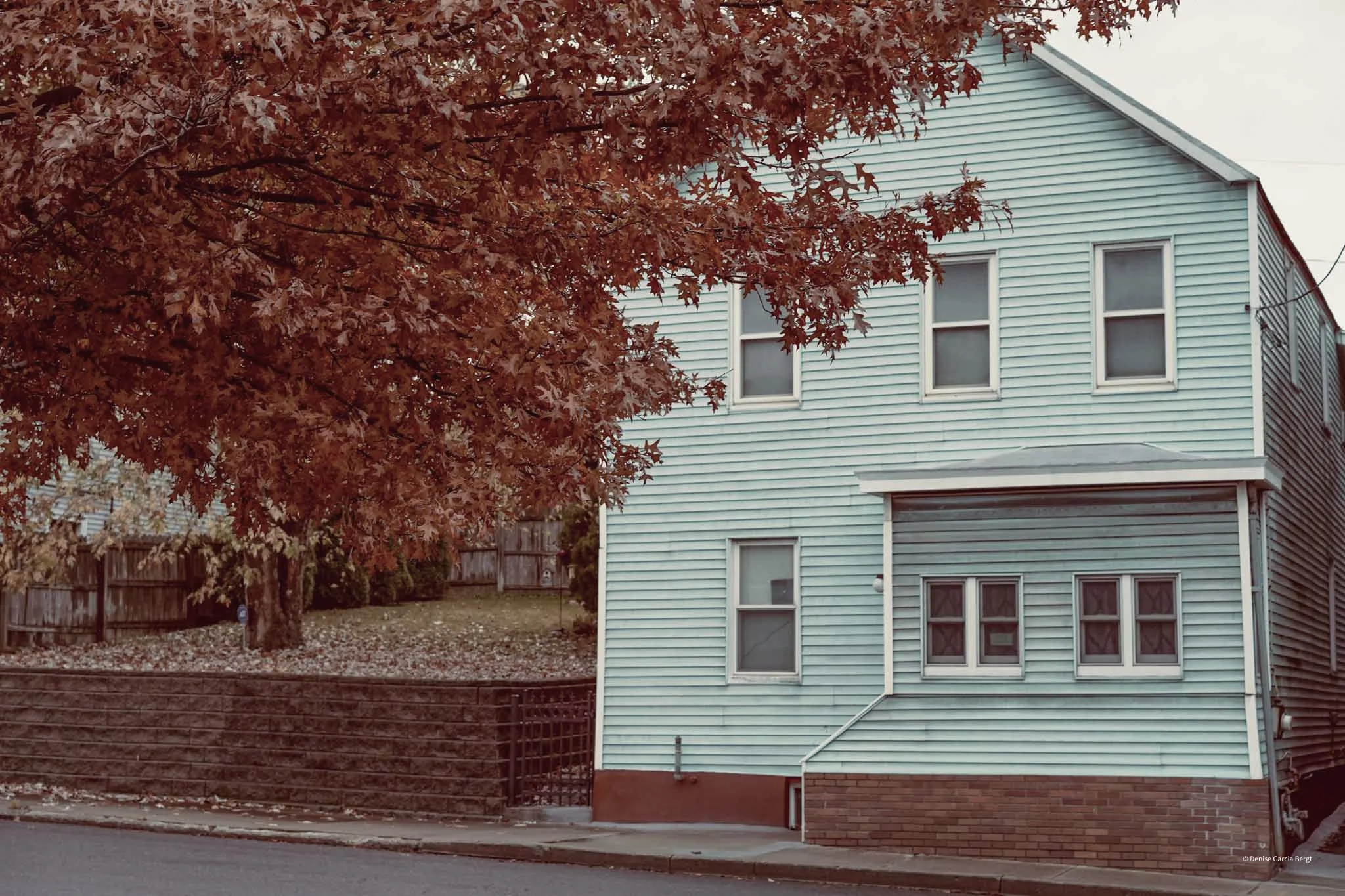 A two-story house with light blue siding, six windows, and a brick foundation. There is a large tree with reddish-brown leaves partially obscuring the house, and a wooden fence surrounds a yard with some fallen leaves.