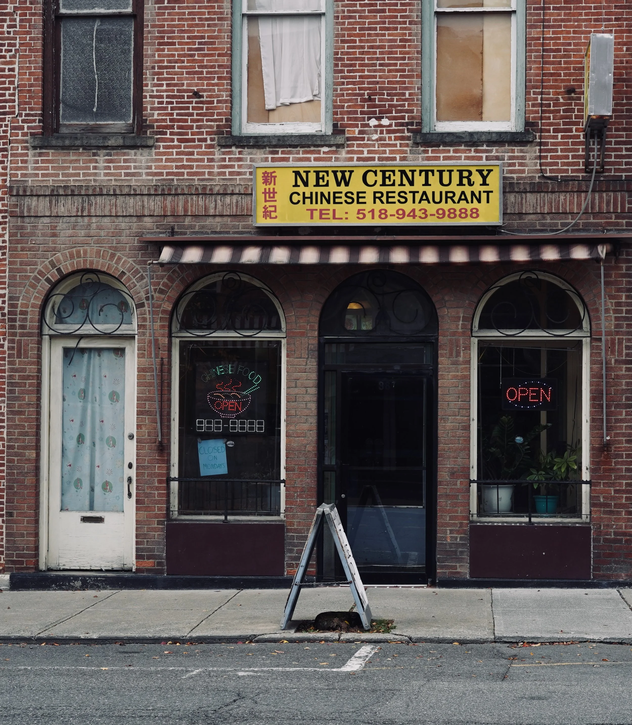 Exterior view of a brick building with a Chinese restaurant named New Century. The restaurant has a yellow sign with black and red lettering, and neon signs in the windows that say "Open" with one also showing a phone number.