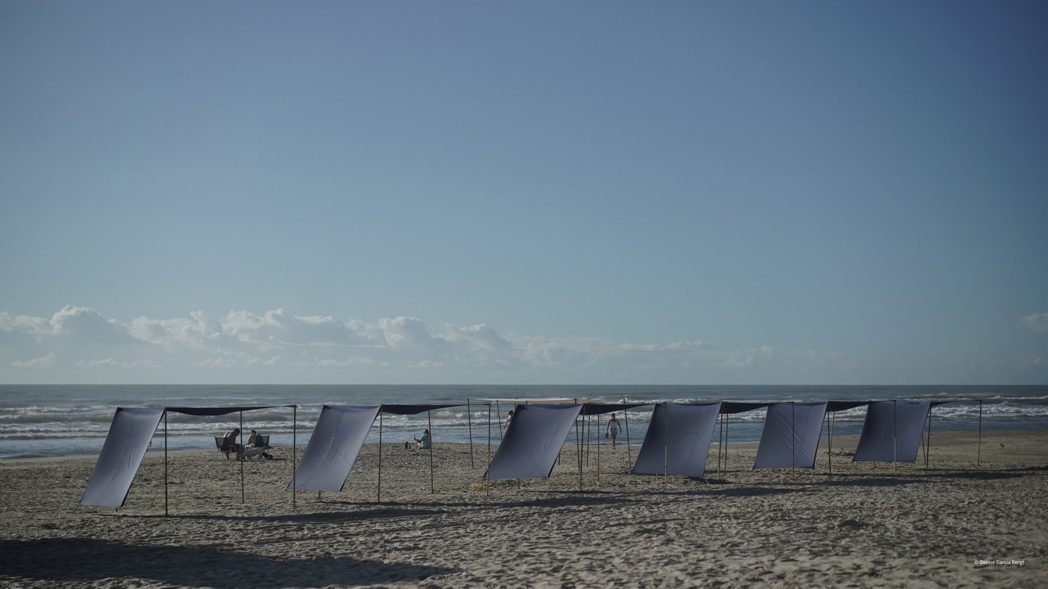 Beach with several windbreaks, a few people sitting and walking near the ocean, and an expansive blue sky with scattered clouds.