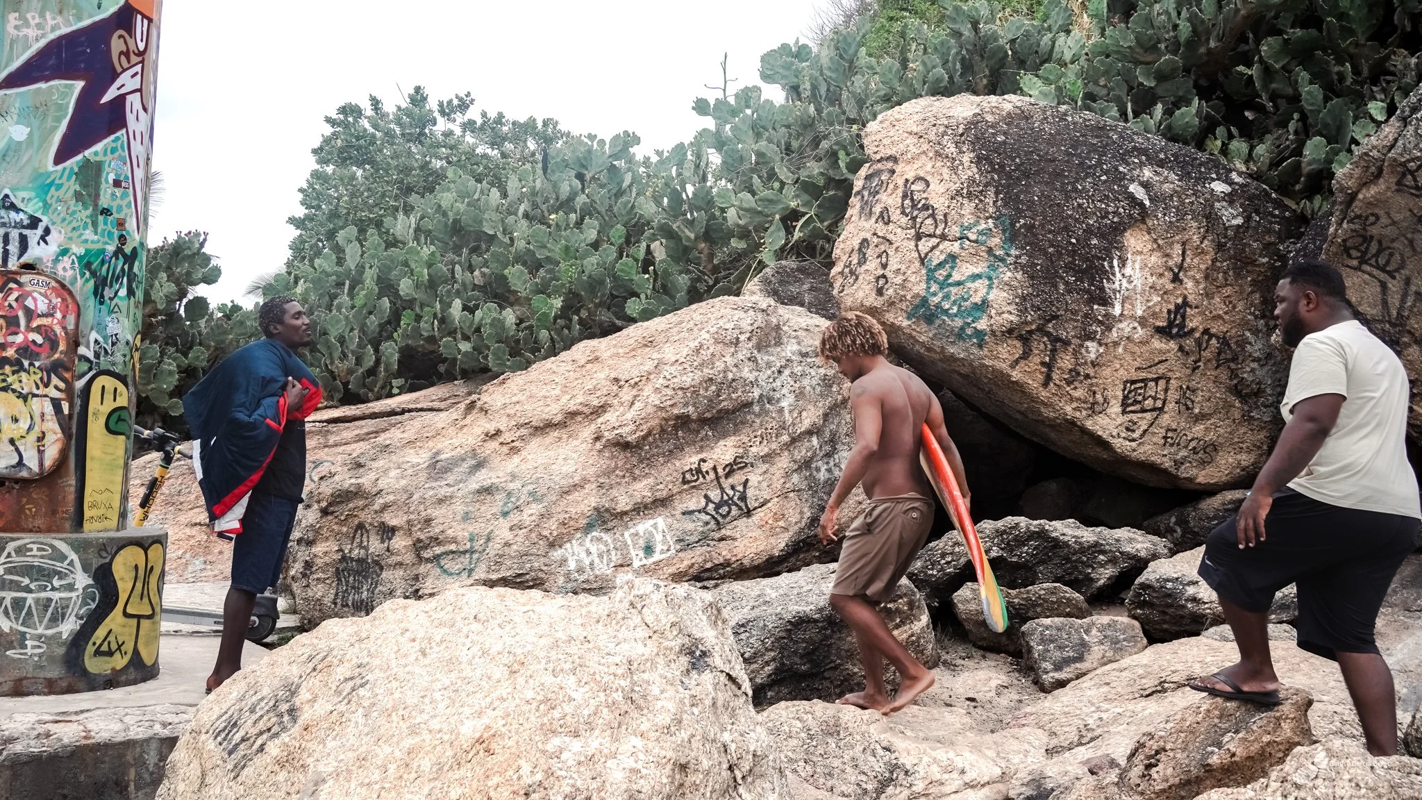 Three young men at a rocky outdoor location with graffiti-covered rocks and cacti in the background. One shirtless with a skateboard, another in a white t-shirt and shorts, and the third holding a backpack and standing near graffiti-covered objects.