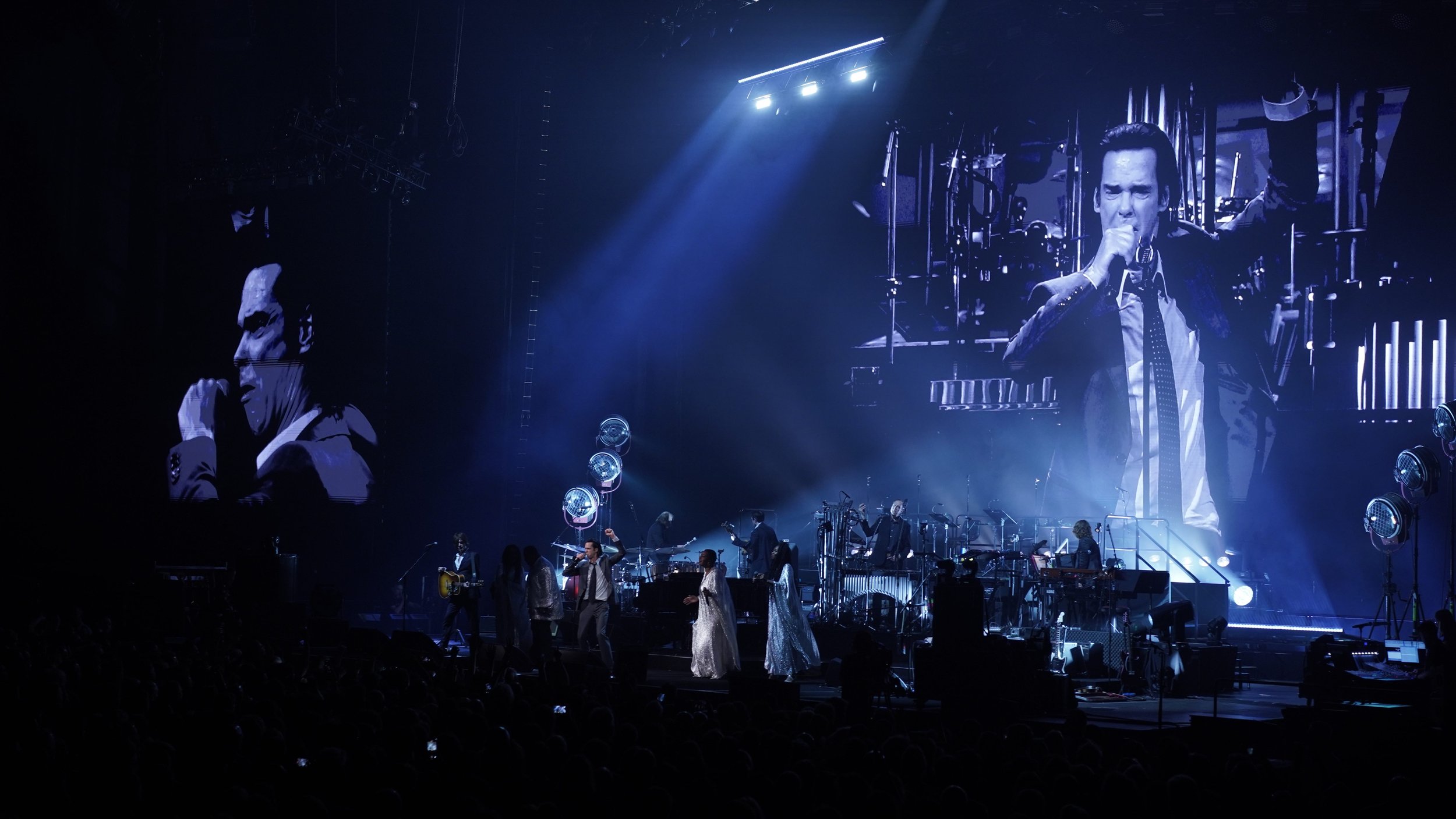 A live concert with musicians on stage, large screens displaying a singer, and dramatic lighting in blue tones.