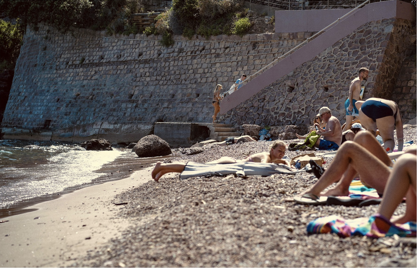People relaxing and sunbathing on a pebbled beach near the water, with a stone wall and stairs leading down to the beach, some people sitting on towels, others walking or standing, wearing swimsuits and beach hats.