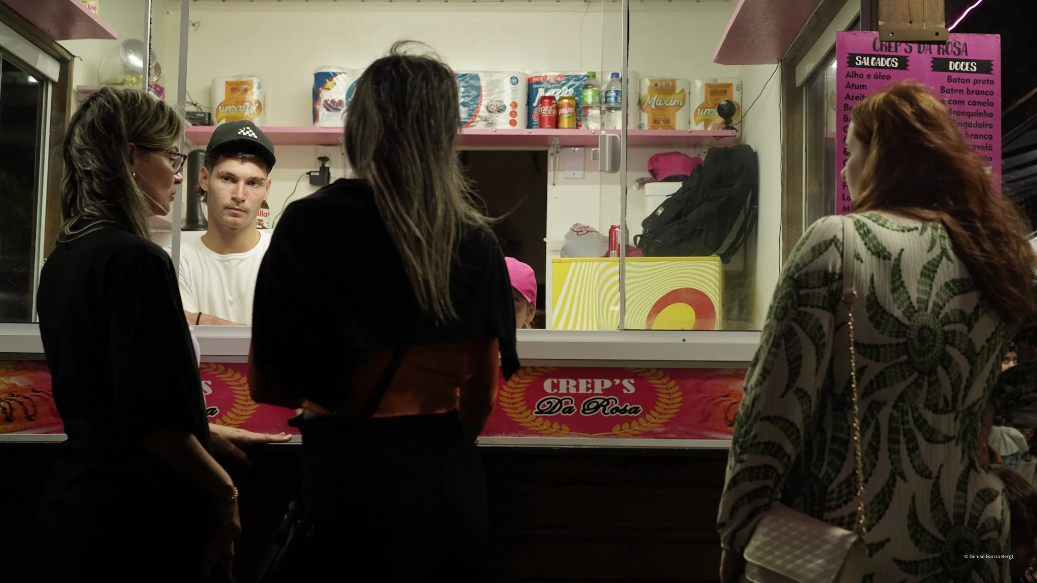 Four people stand in front of a food cart named 'Crep's D'a Rosa,' which sells crepes. Two women and one man are visible on one side of the window, and a woman with long hair on the other side, behind the counter, preparing food. The cart has various