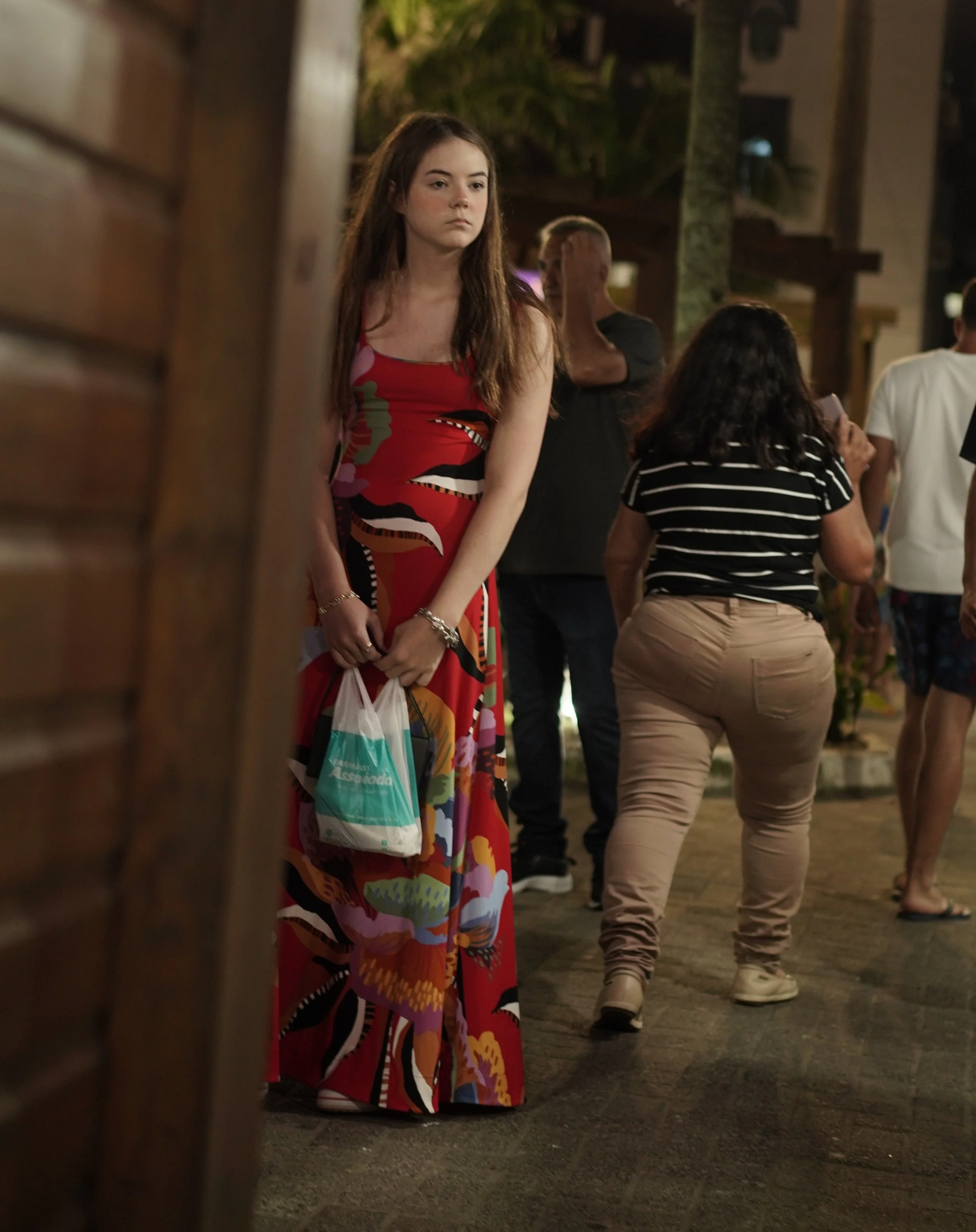 A young woman in a colorful long dress holding a plastic bag standing outdoors at night, surrounded by other people.