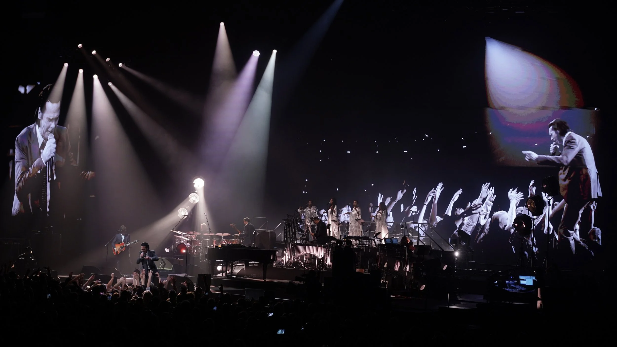 A concert stage with a singer holding a microphone, a guitarist, and a group of backup singers, with large screens displaying the performers and an enthusiastic audience.