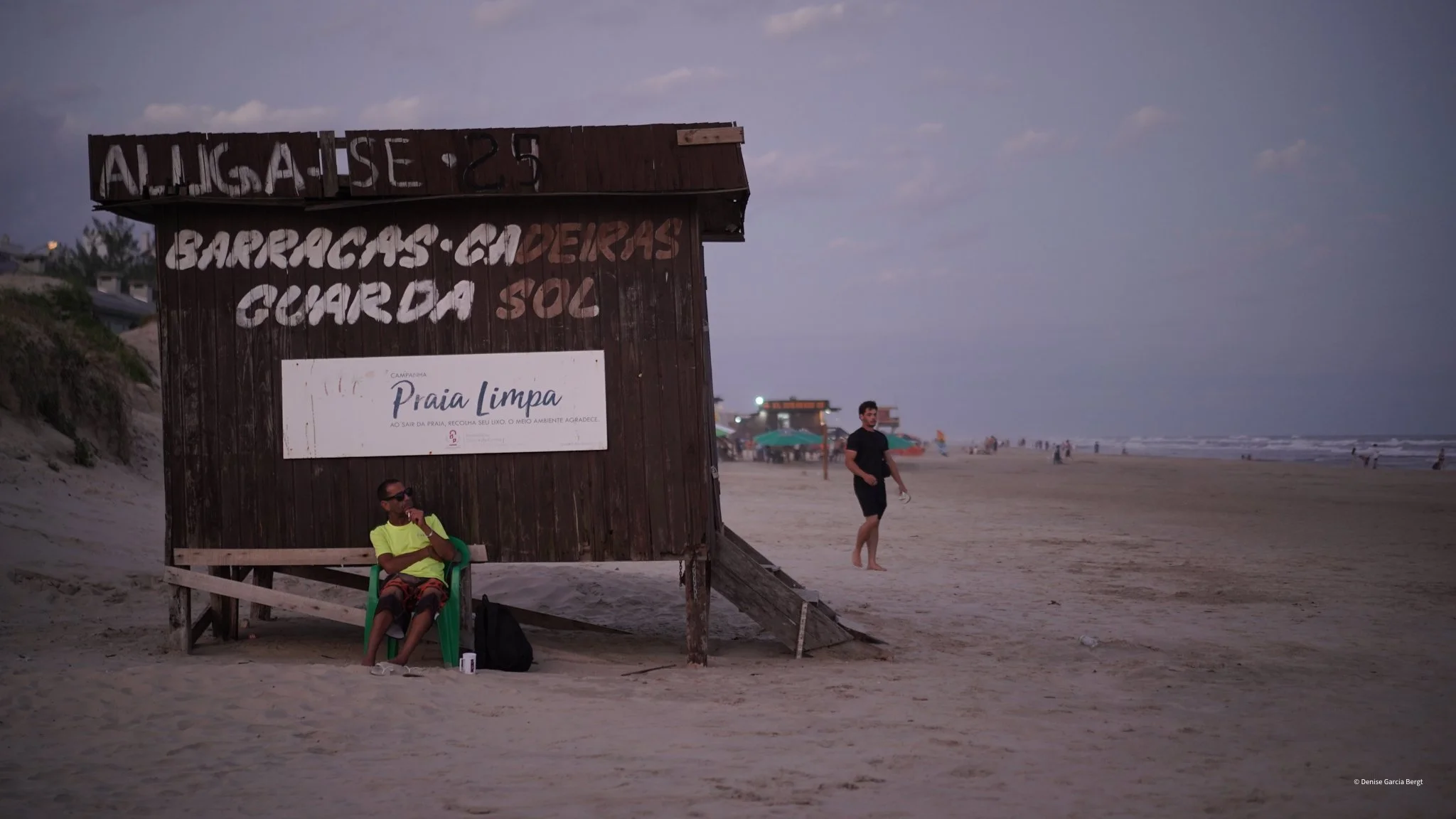 A beach scene during dusk with a wooden lifeguard stand featuring a sign, a man sitting on a green chair with a drink, and another man walking along the shore with umbrellas and beachgoers in the background.