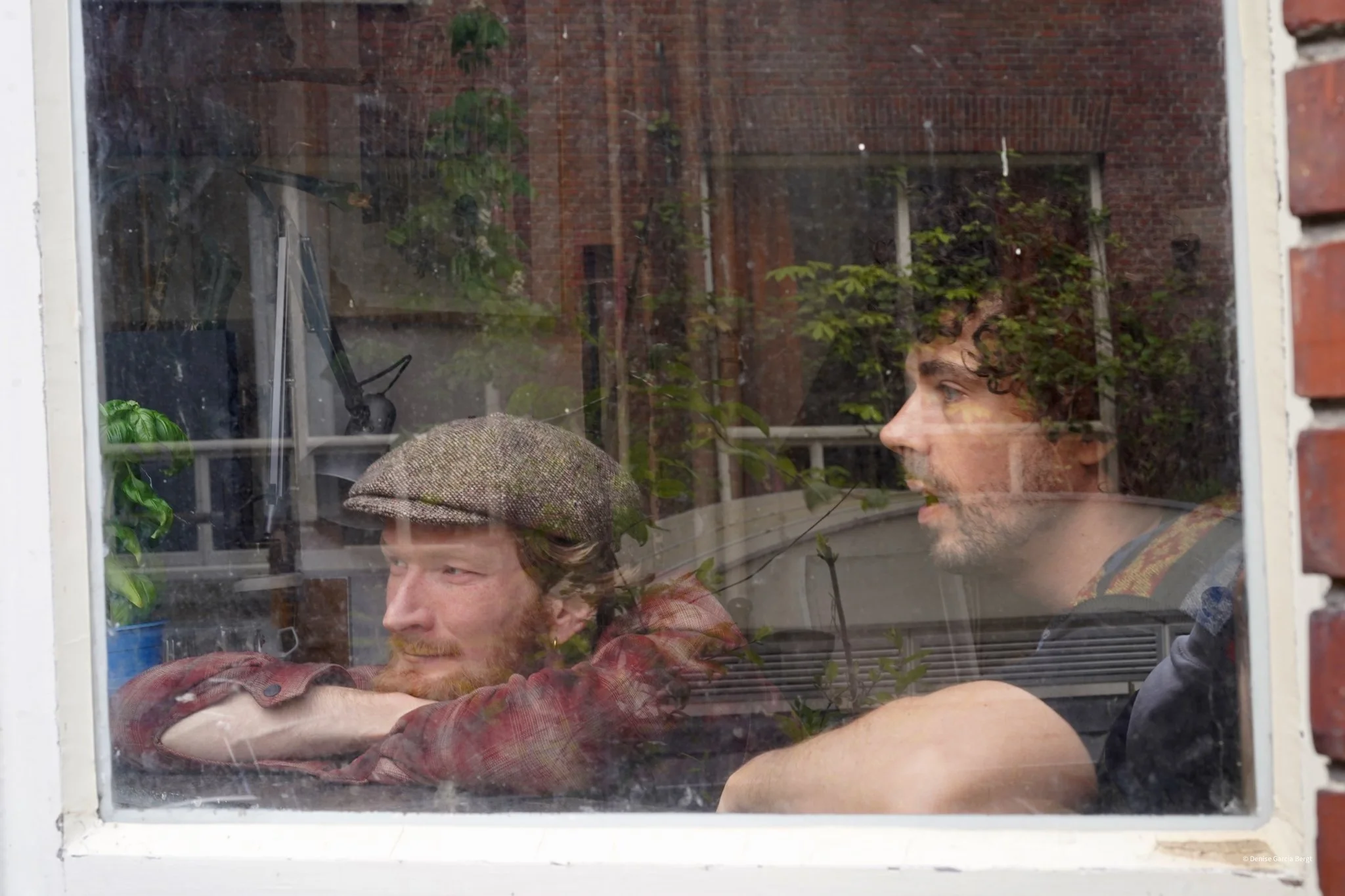 Two men sitting inside, seen through a window with reflections. One man with a beard wears a plaid shirt and a flat cap, while the other with curly hair and beard wears a dark shirt.