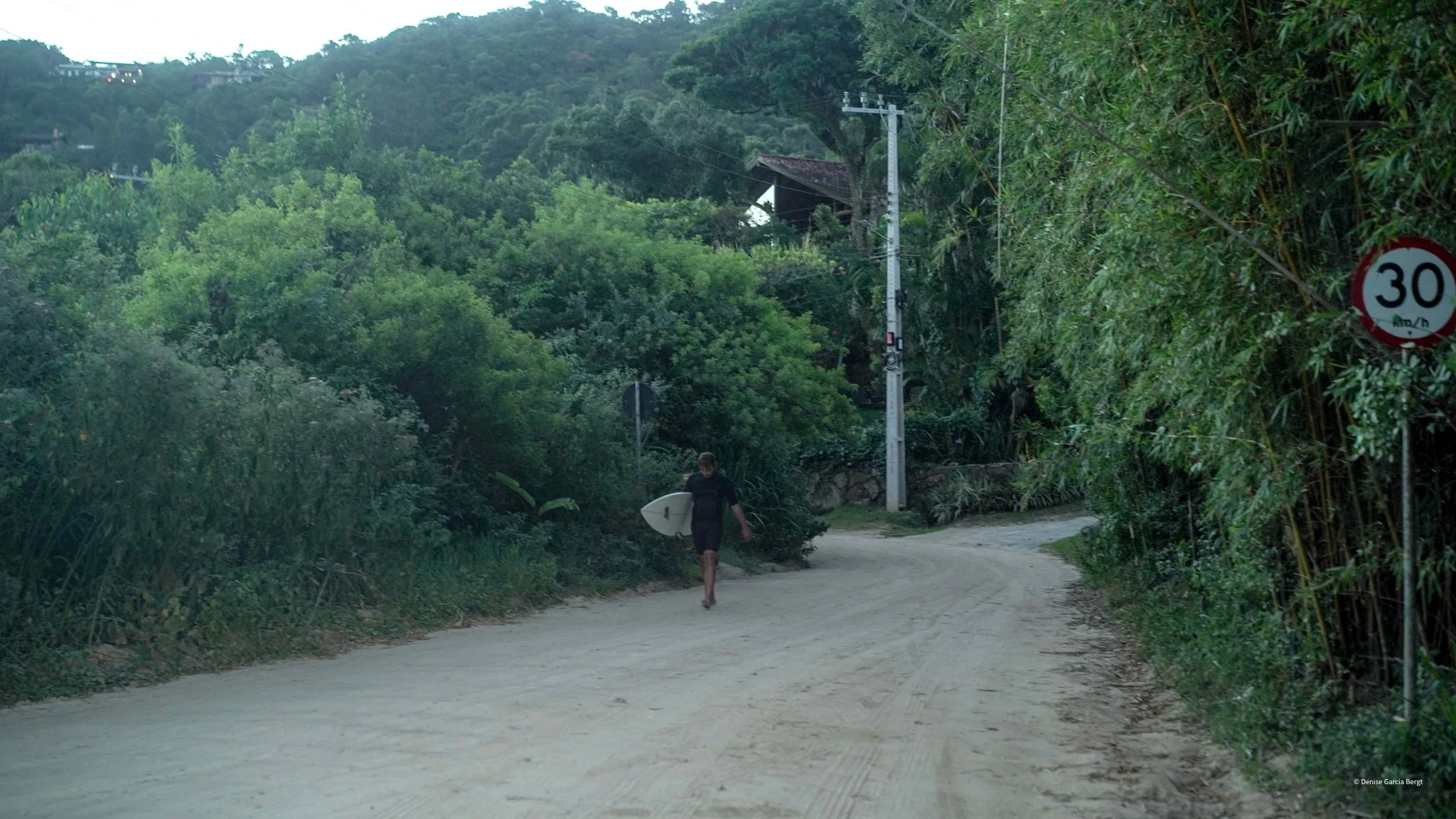 A person walking down a dirt road carrying a surfboard, surrounded by lush green trees and bushes, with utility poles and a 30 km/h speed limit sign on the right.