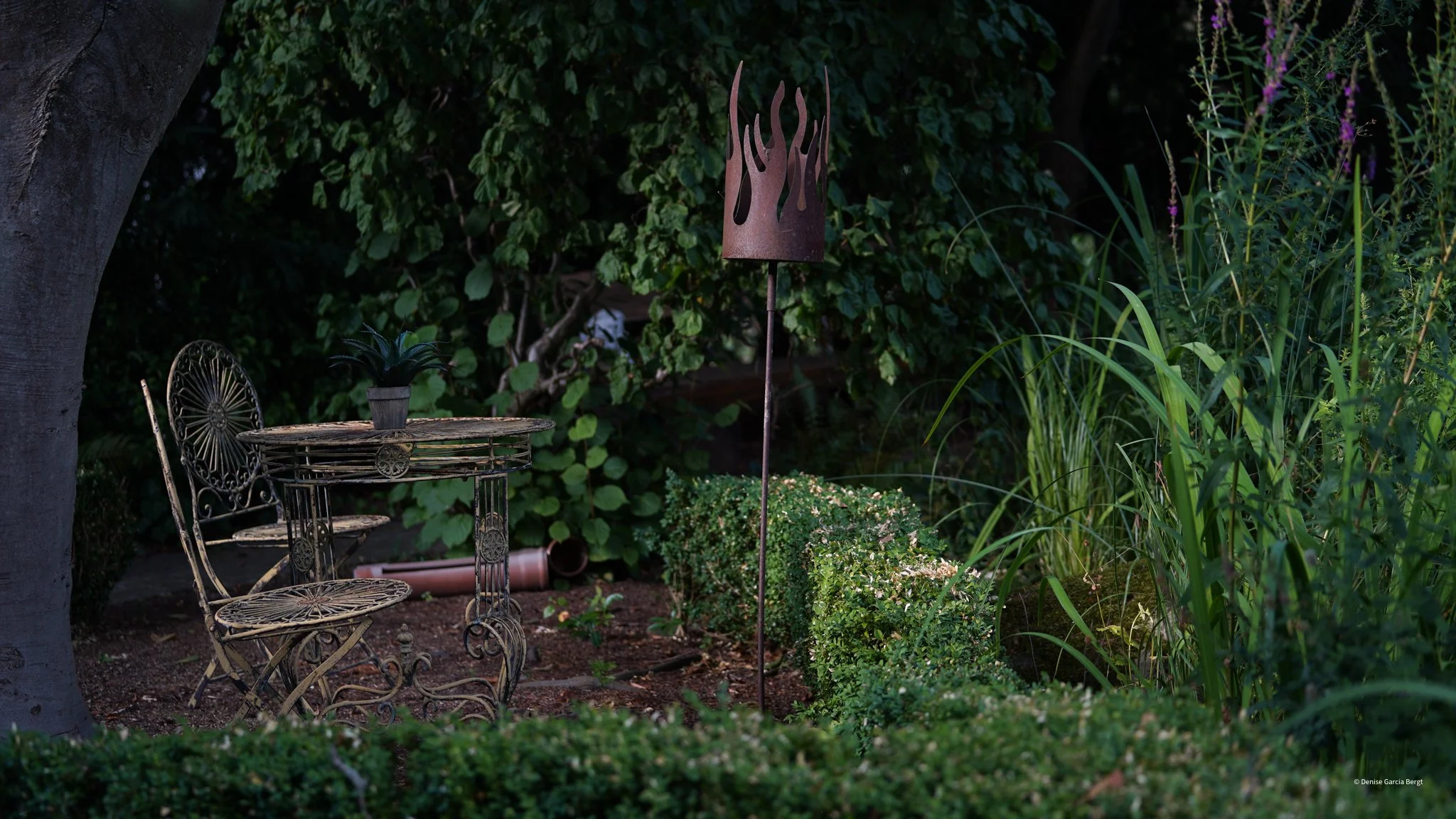 A garden scene featuring a small round metal table with a potted plant on top, a matching metal chair, surrounded by lush green plants, bushes, and trees, with a metal flame-shaped garden ornament in the background.
