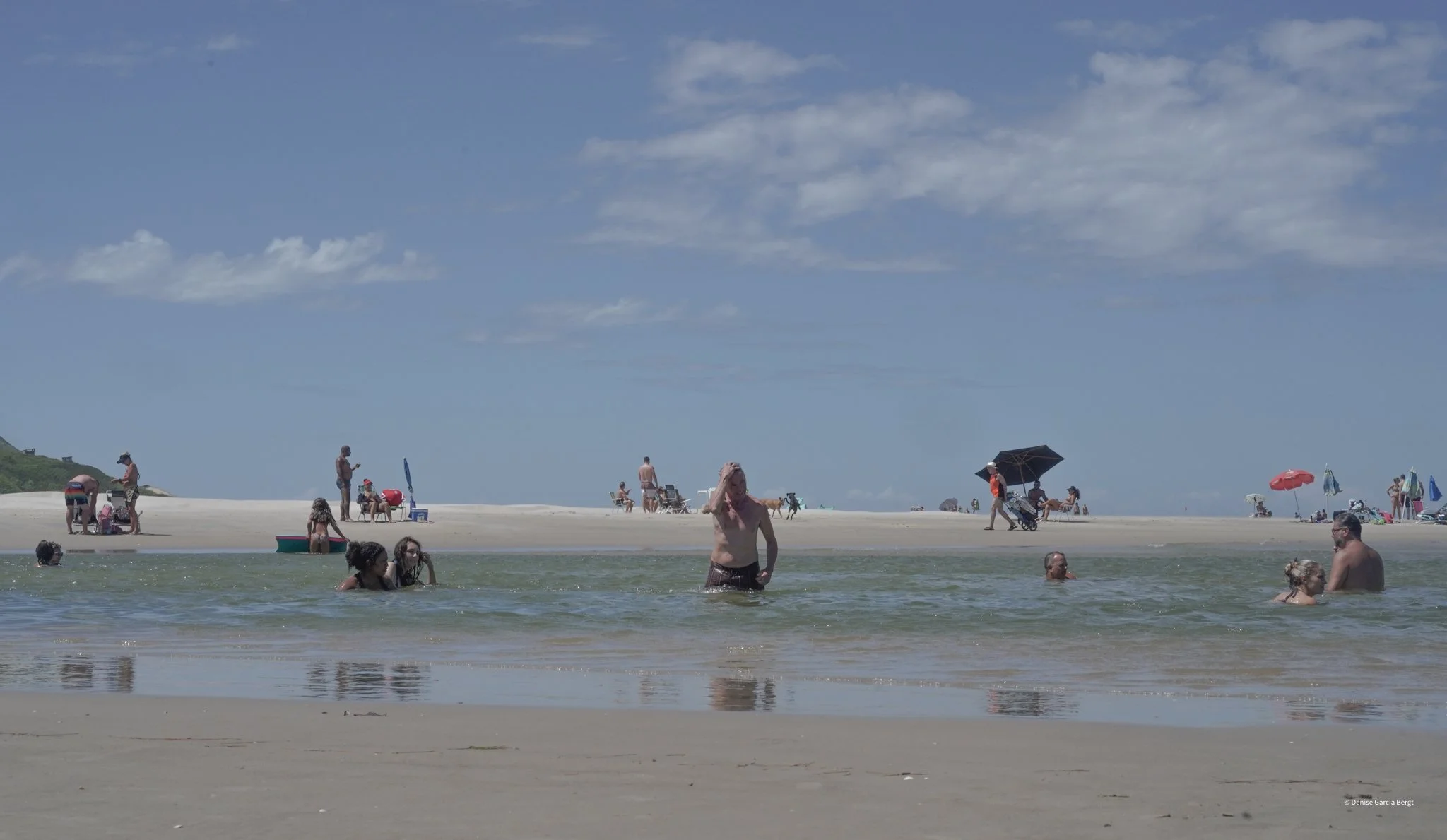 People enjoying a day at the beach; some are swimming in the water, others are sitting or walking on the sandy shore under a partly cloudy sky.