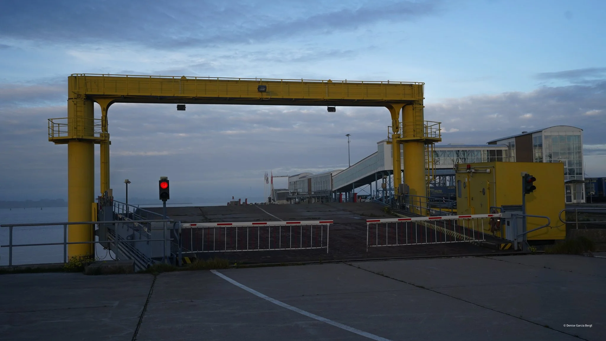 A closed ferry dock with a red traffic light in front, a yellow crane structure, and a building in the background on a cloudy day.