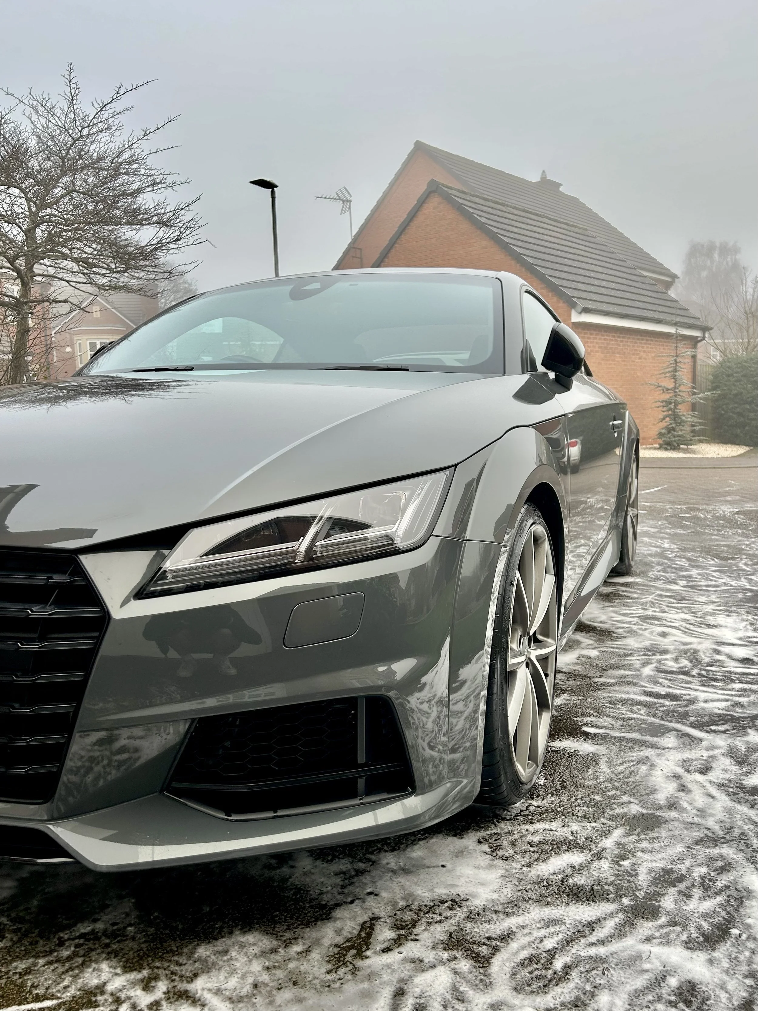 Gray sports car parked on a snow-covered driveway in front of a brick house on a foggy day.