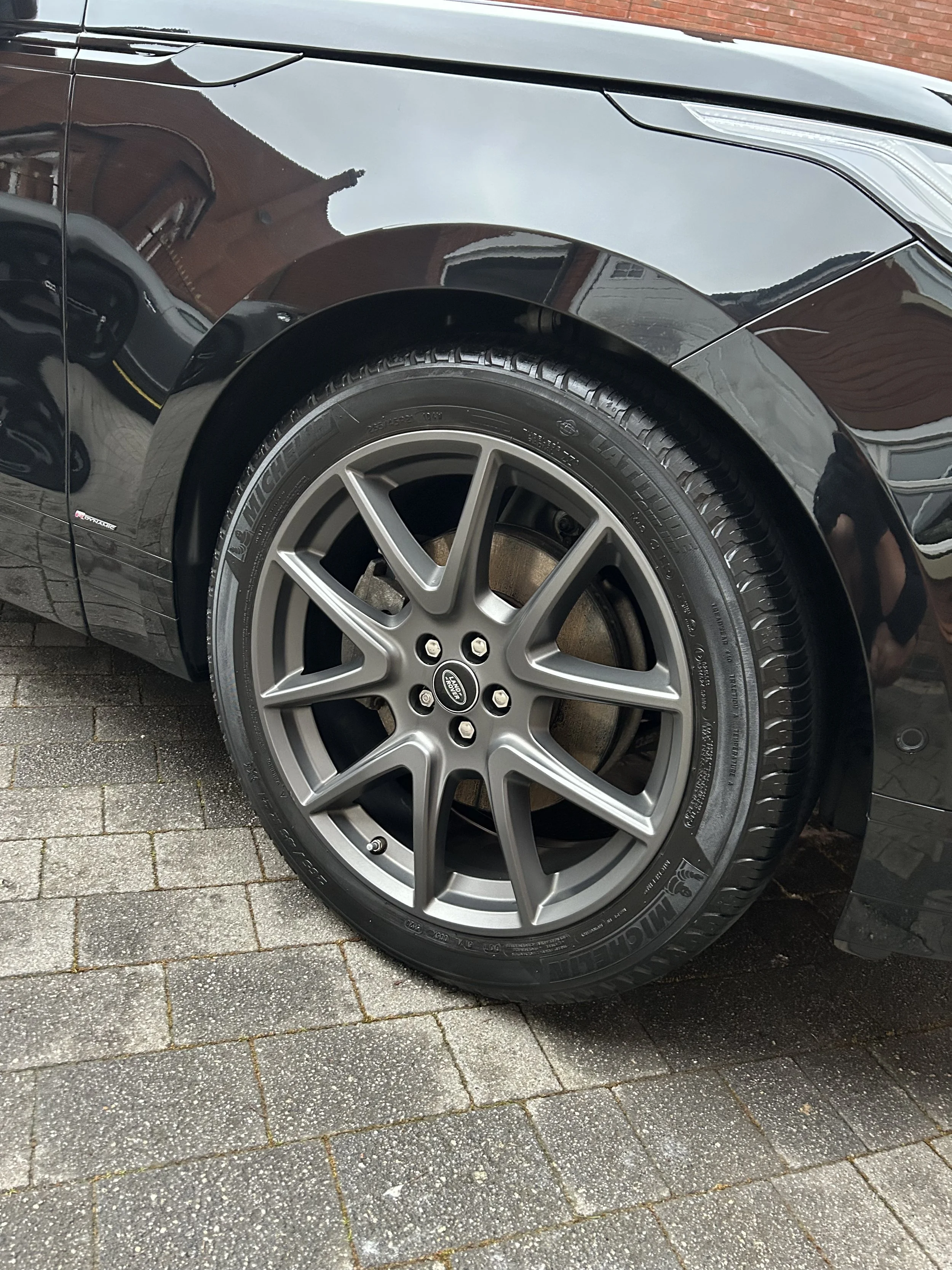 Close-up of a black car's front tire and wheel, showing a multi-spoke wheel and Michelin tire, parked on a brick-paved driveway.